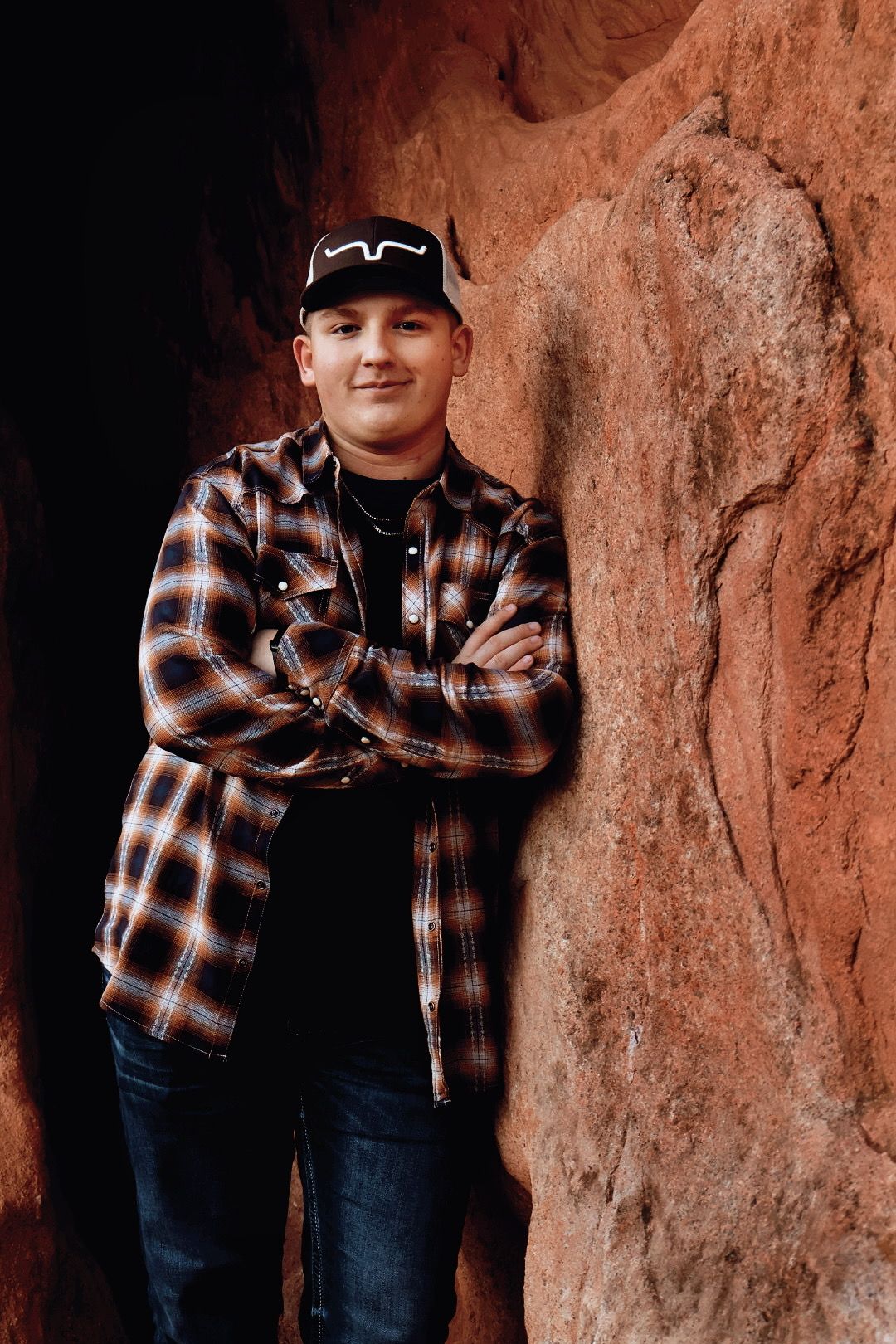Senior boy in flannel leans confidently on red rock wall. Outdoor senior portrait in Colorado Springs.