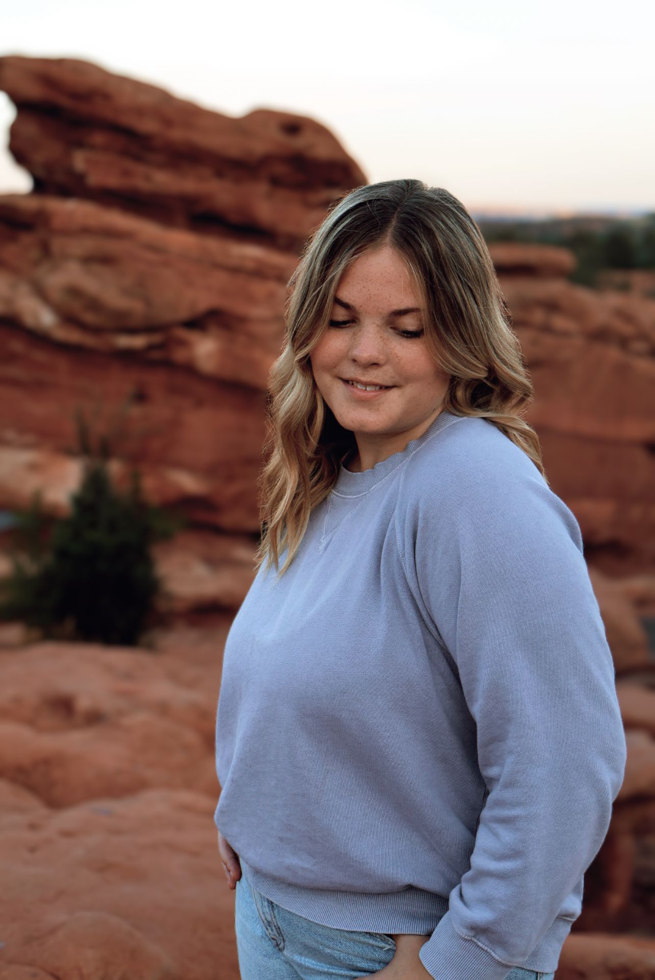 Blonde girl in purple sweatshirt smiles shyly with red rocks behind her. Sunset senior photography session.