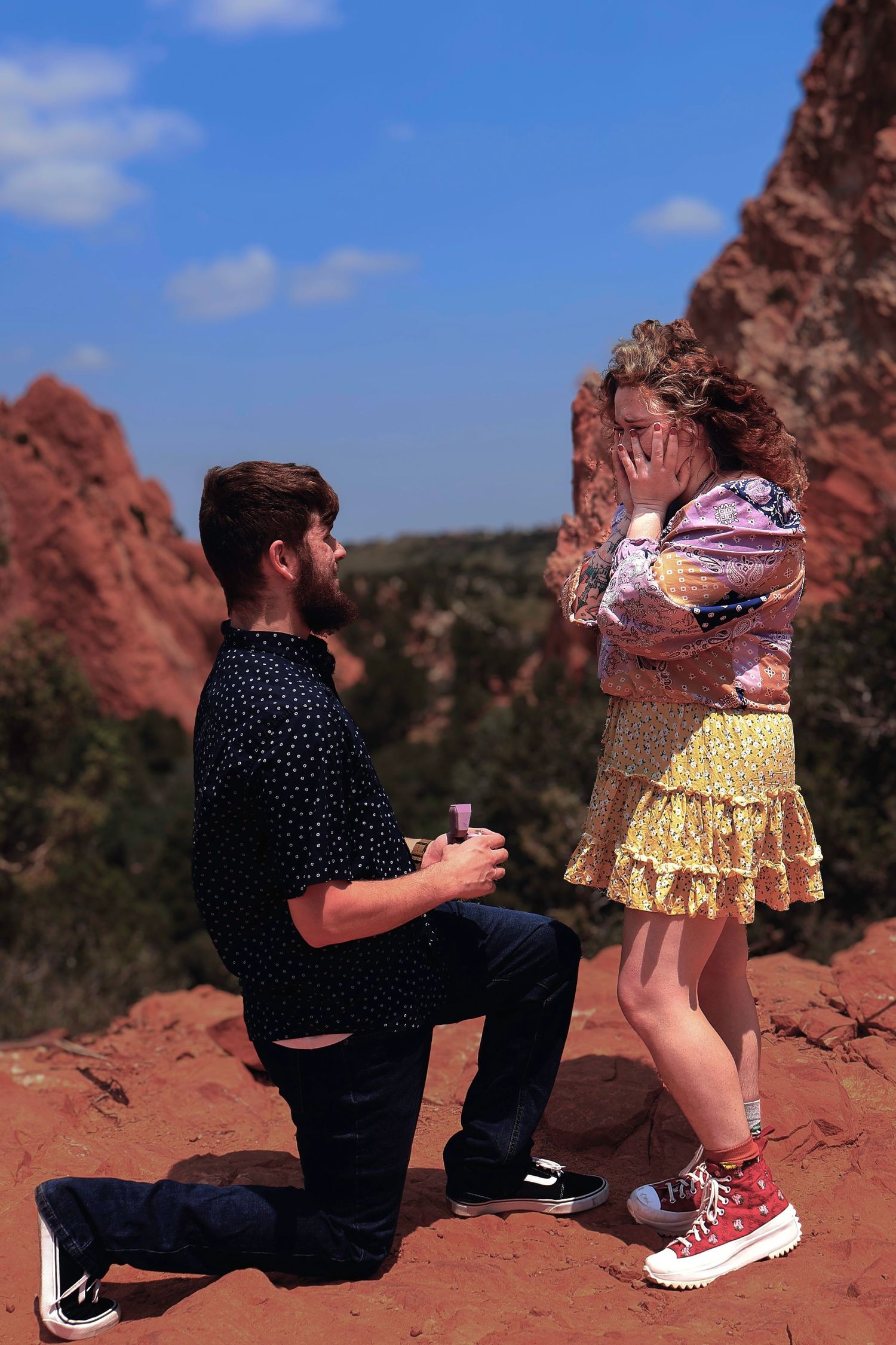 Man proposes on one knee to surprised woman covering her face among red rock formations under blue sky.