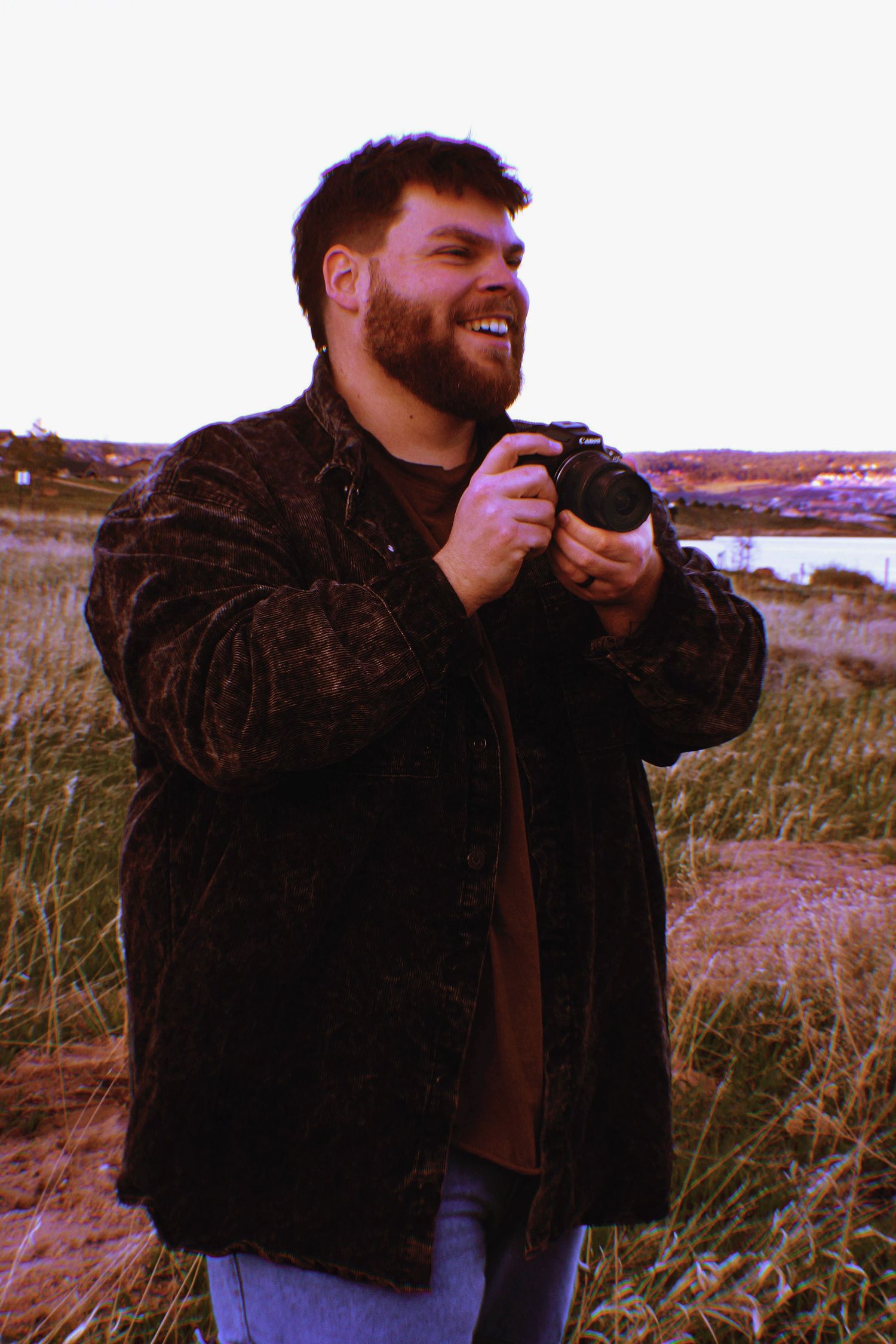 Man with a camera in hand smiling joyfully, standing in tall grass at golden hour with blurred landscape in the background.