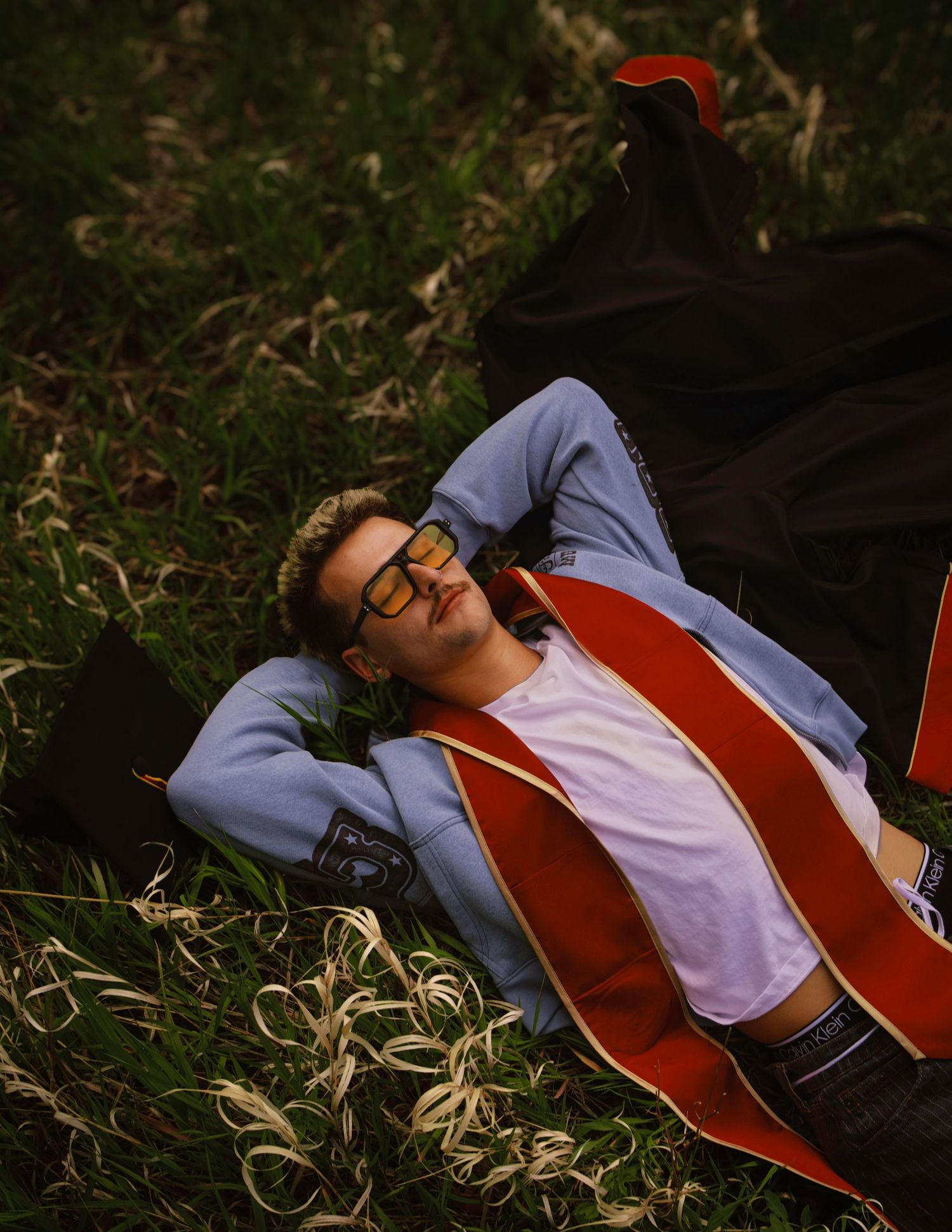 Young man in graduation gown lying on grass, wearing glasses and a red sash, relaxing outdoors.