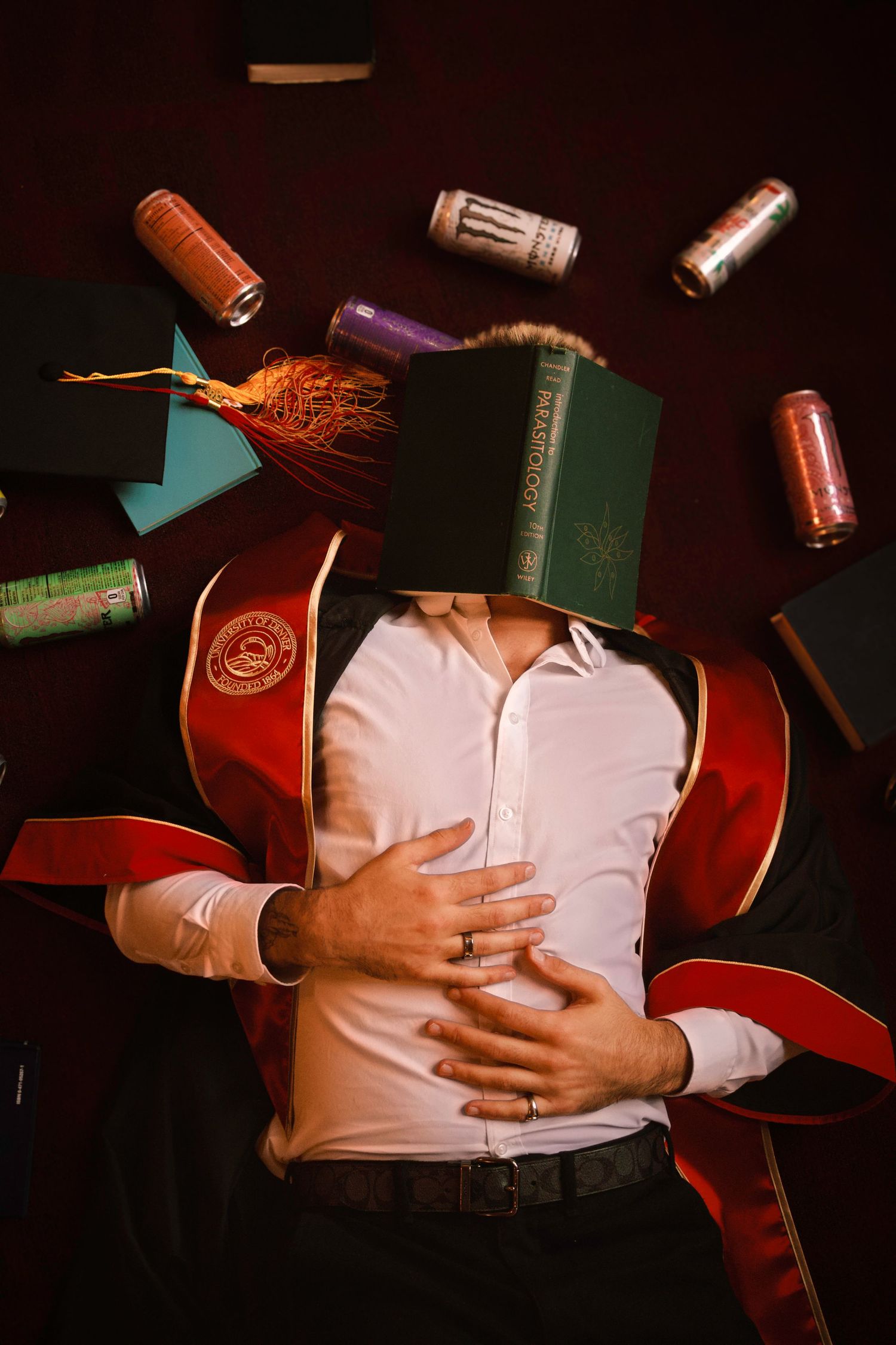 Colorado Springs Graduate in red robe lying down surrounded by flying books and energy drinks in a dramatic dark setting.