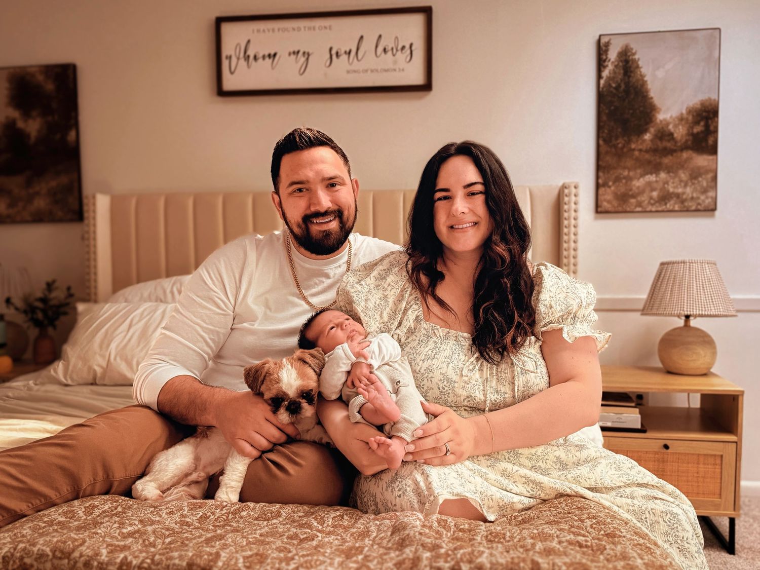 A happy couple sits on a bed with a small dog and newborn baby in a cozy, warmly decorated bedroom.