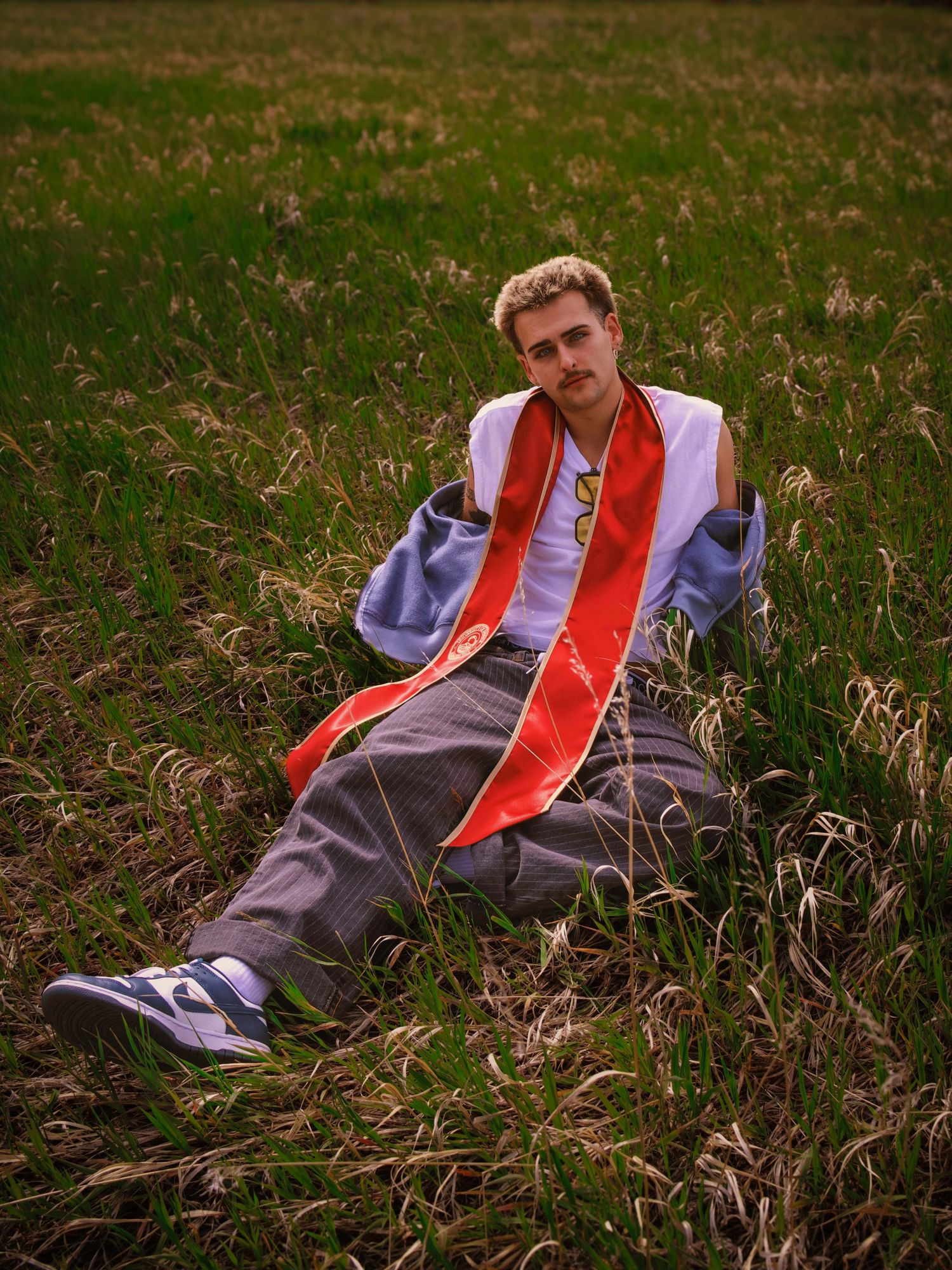 Young man in graduation sash sits relaxed in a lush green field wearing sneakers and casual gray pants.
