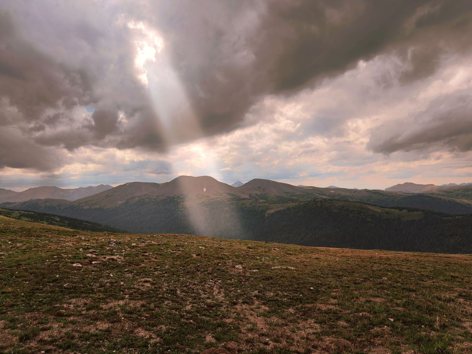 Dramatic sunbeam breaking through storm clouds over a scenic mountain landscape with rolling green hills in Rocky Mountain National Park.