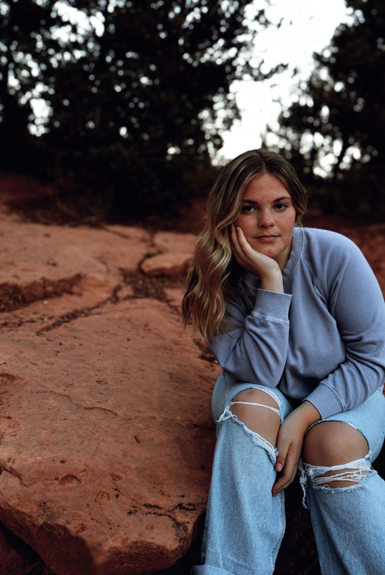 Teen girl sits on red rock trail with hand on cheek, soft smile. Natural light senior portrait in Colorado.