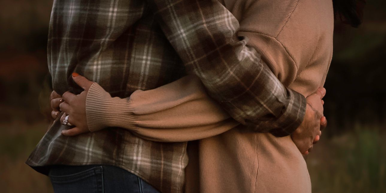 Couple embracing outdoors, wearing plaid and tan coats, sharing a warm hug in a natural setting.