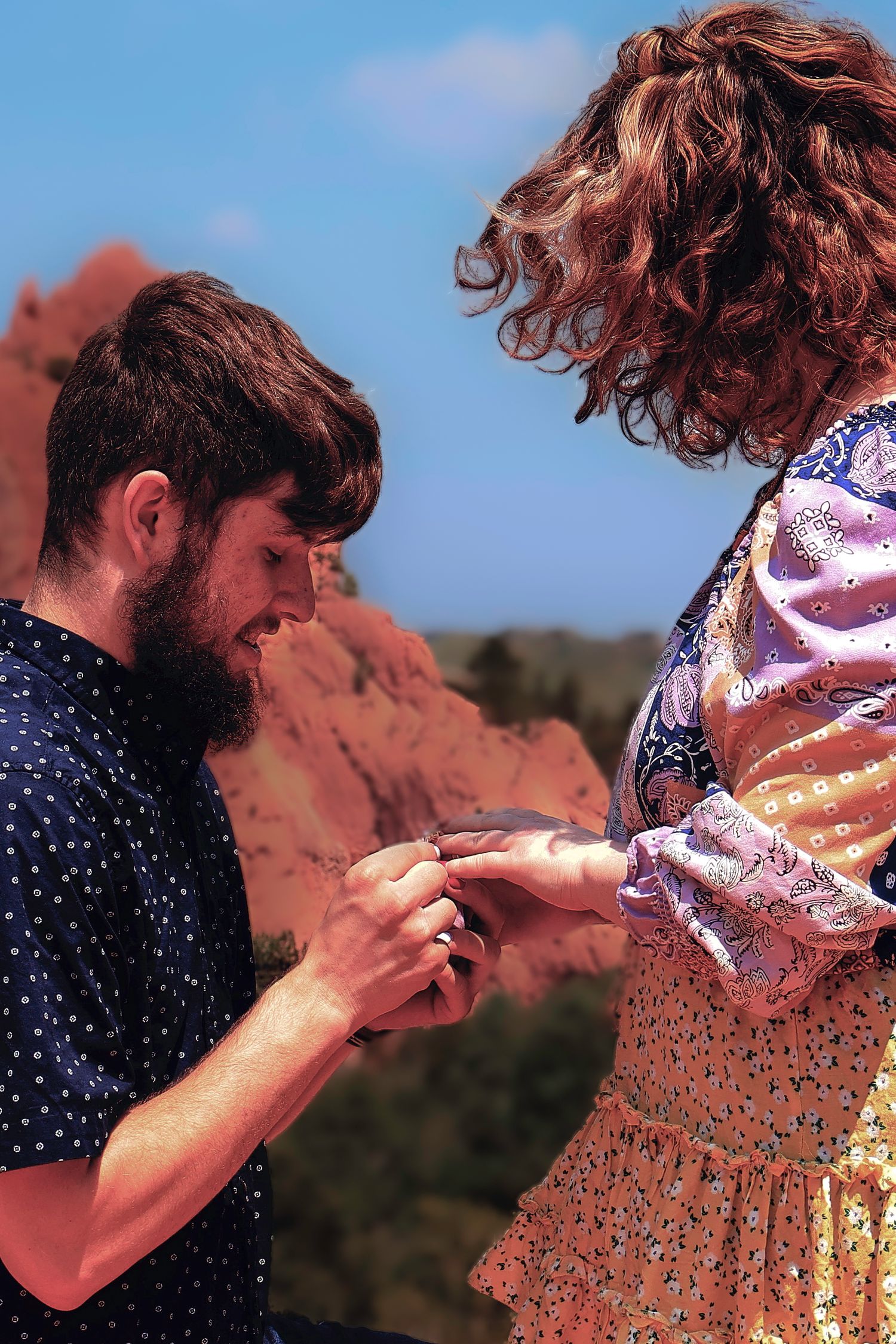 Man kneeling to propose to woman outdoors near red rock formations under a clear blue sky.
