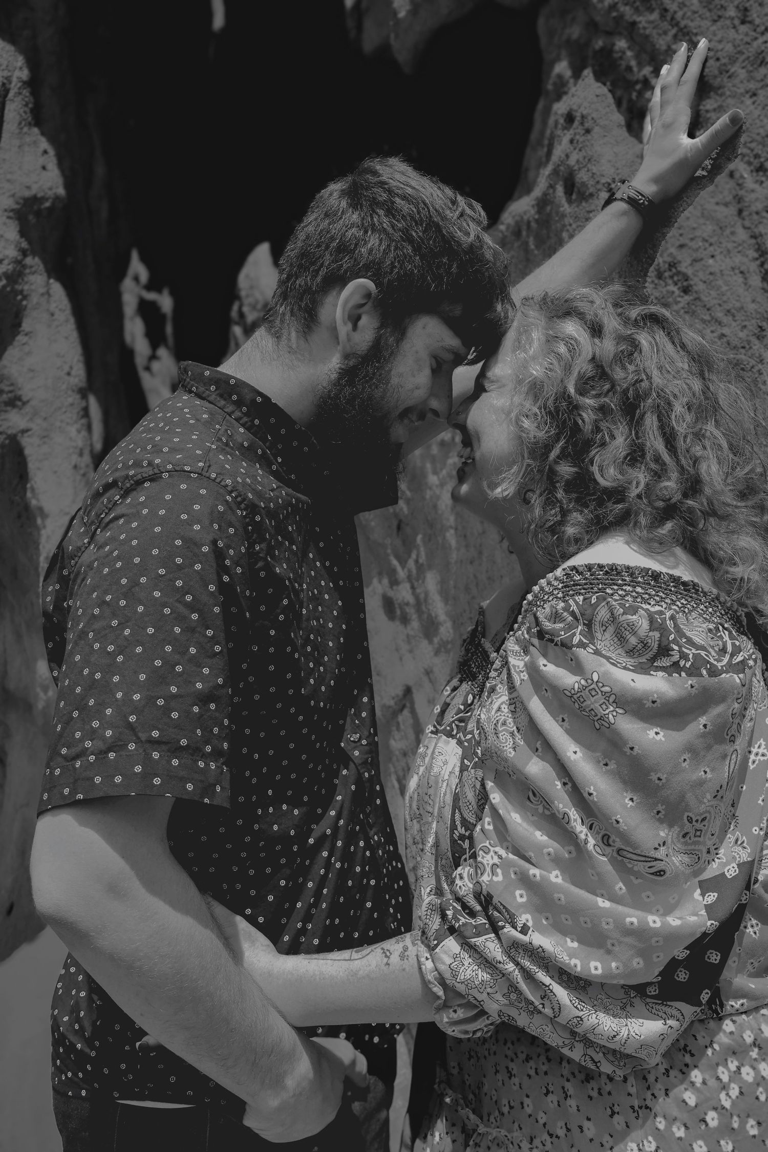 Black and white photo of a couple sharing a romantic moment, leaning in close to kiss near a rocky wall.