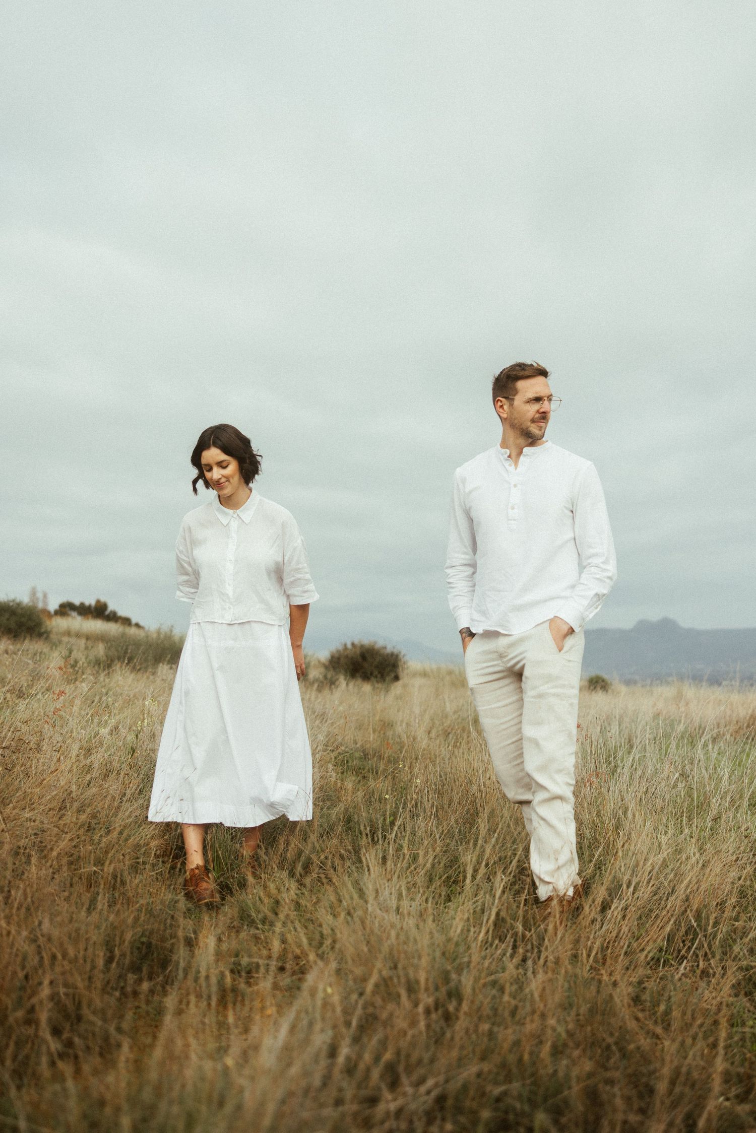 Couple in white clothing walking through a field on an overcast day.