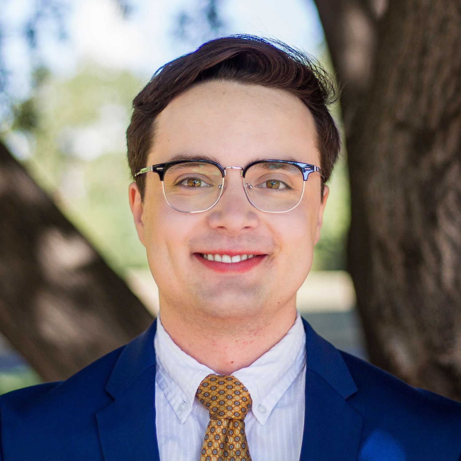 Man in business attire smiling while wearing glasses and a tie against outdoor background.