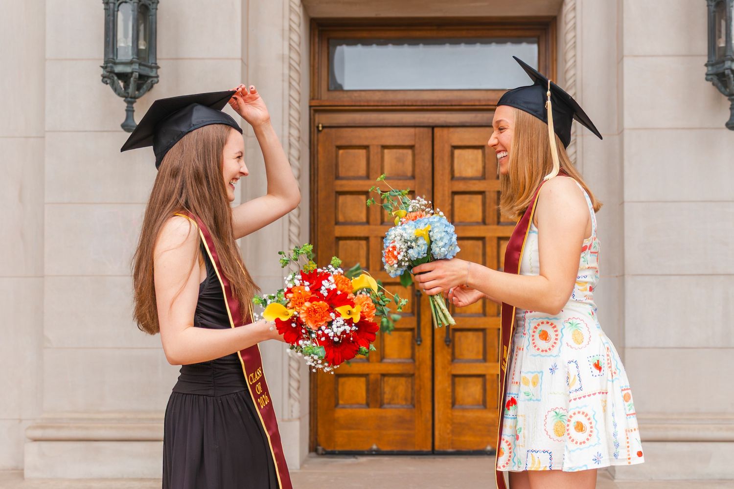 Two female graduates in black caps holding colorful bouquets and laughing together outside wooden doors.