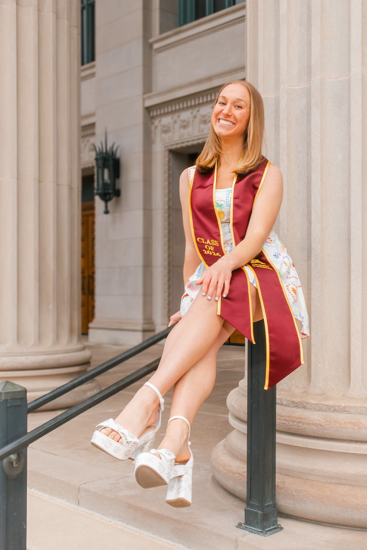 Smiling graduate in maroon cap and gown sits on steps of columned building wearing white platform sandals.
