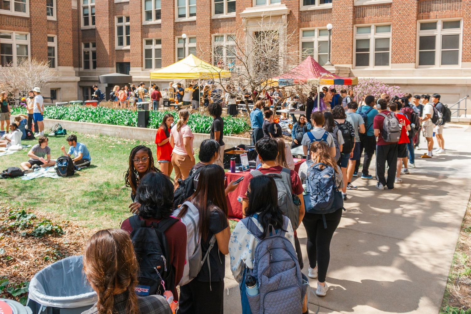 Crowded college campus courtyard with students walking and gathering near brick buildings on a sunny day.