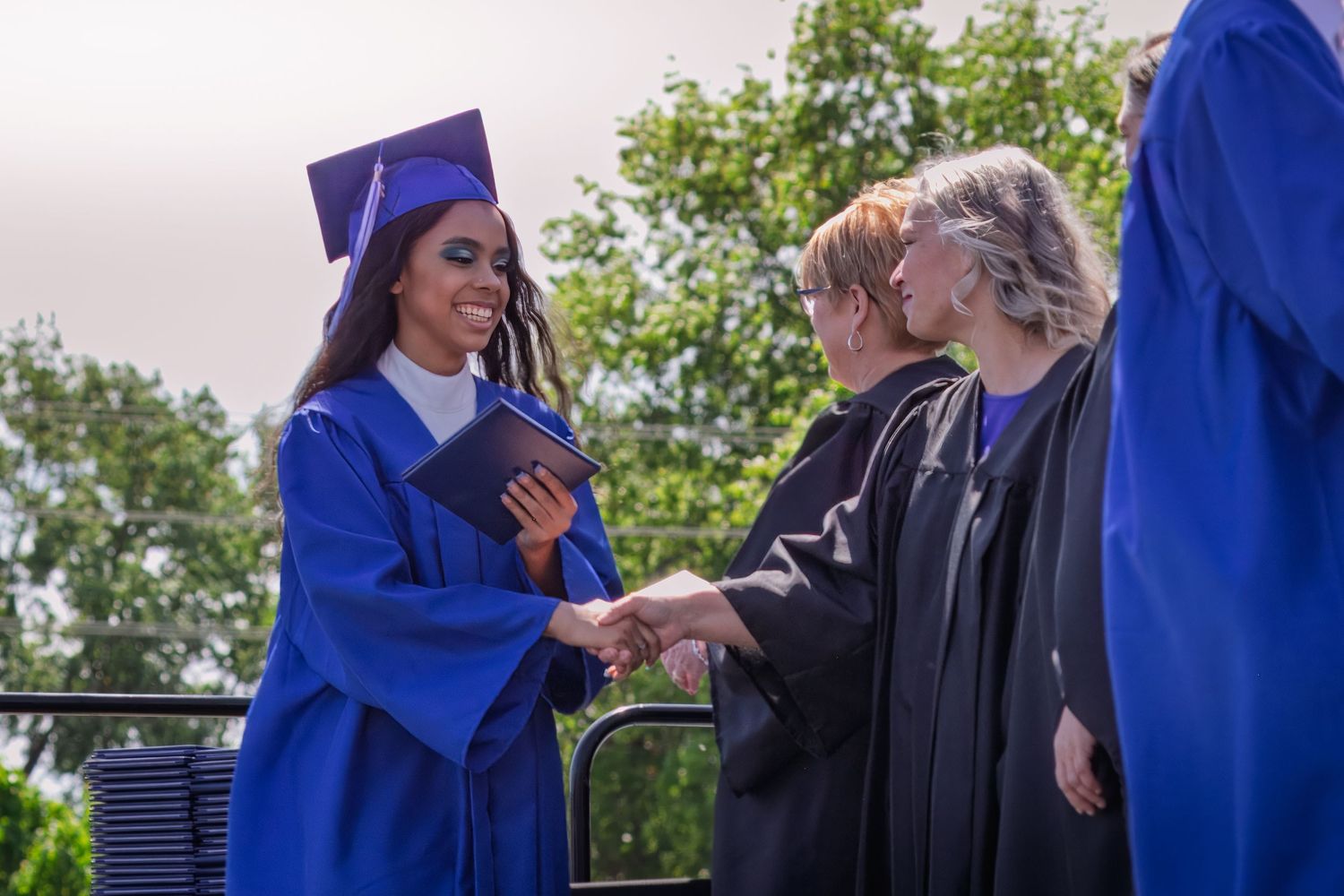 A graduate in royal blue regalia receives their diploma while shaking hands with faculty during outdoor graduation.
