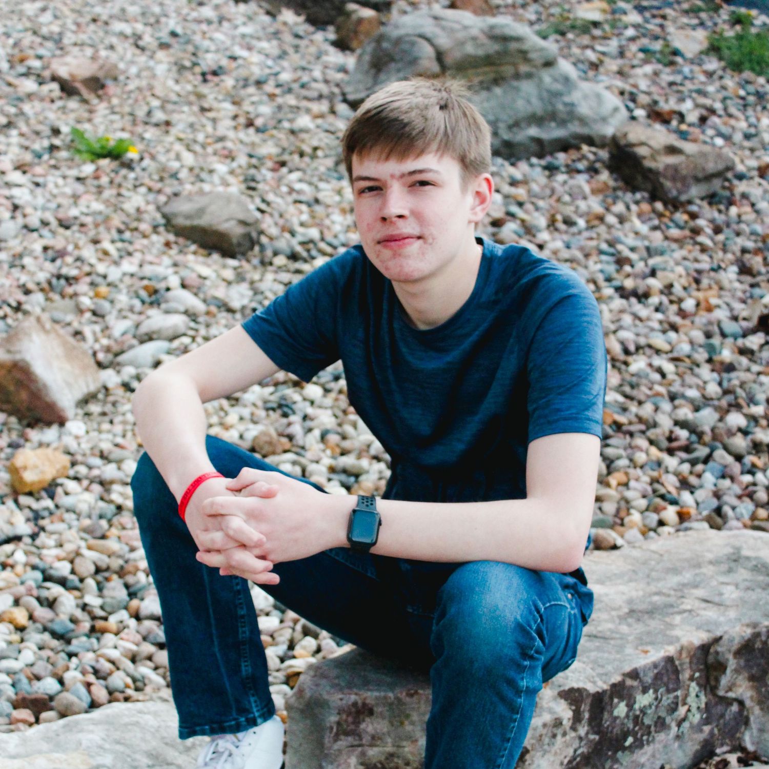 Teen in blue t-shirt sitting on rocky beach looking at camera.