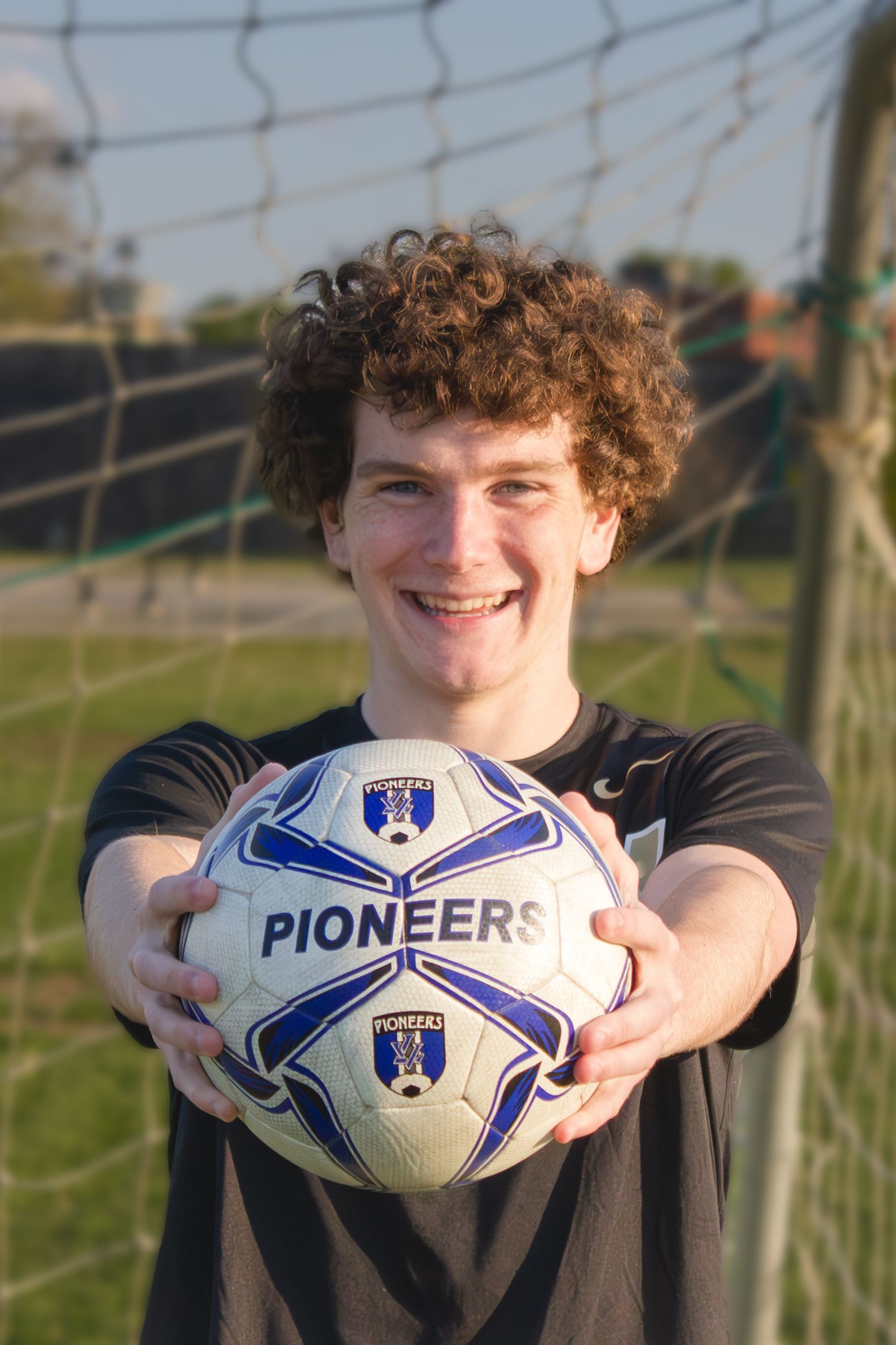 Person wearing a black shirt holds a Pioneers soccer ball in front of a goal net.