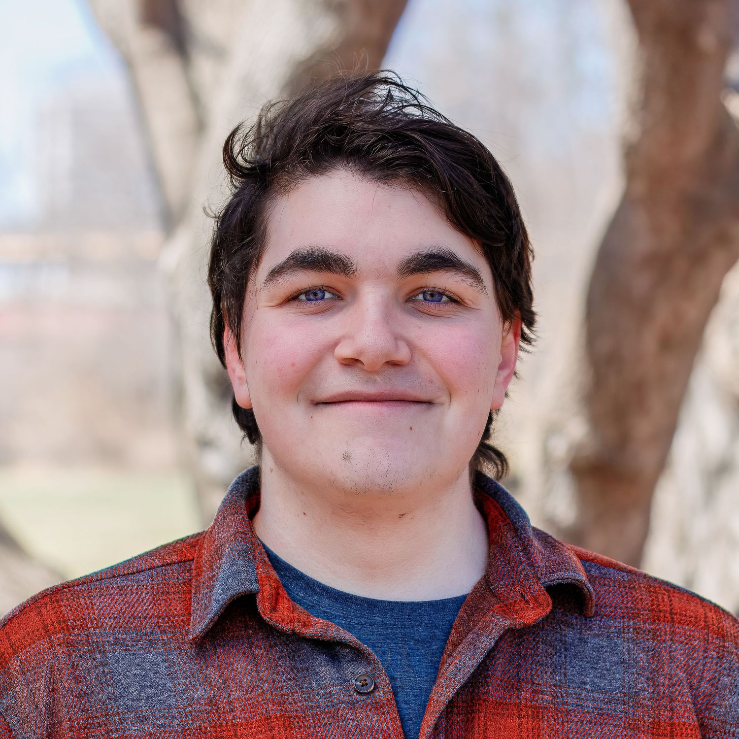 A portrait shot of a person wearing a red plaid flannel shirt and blue t-shirt while standing outdoors.