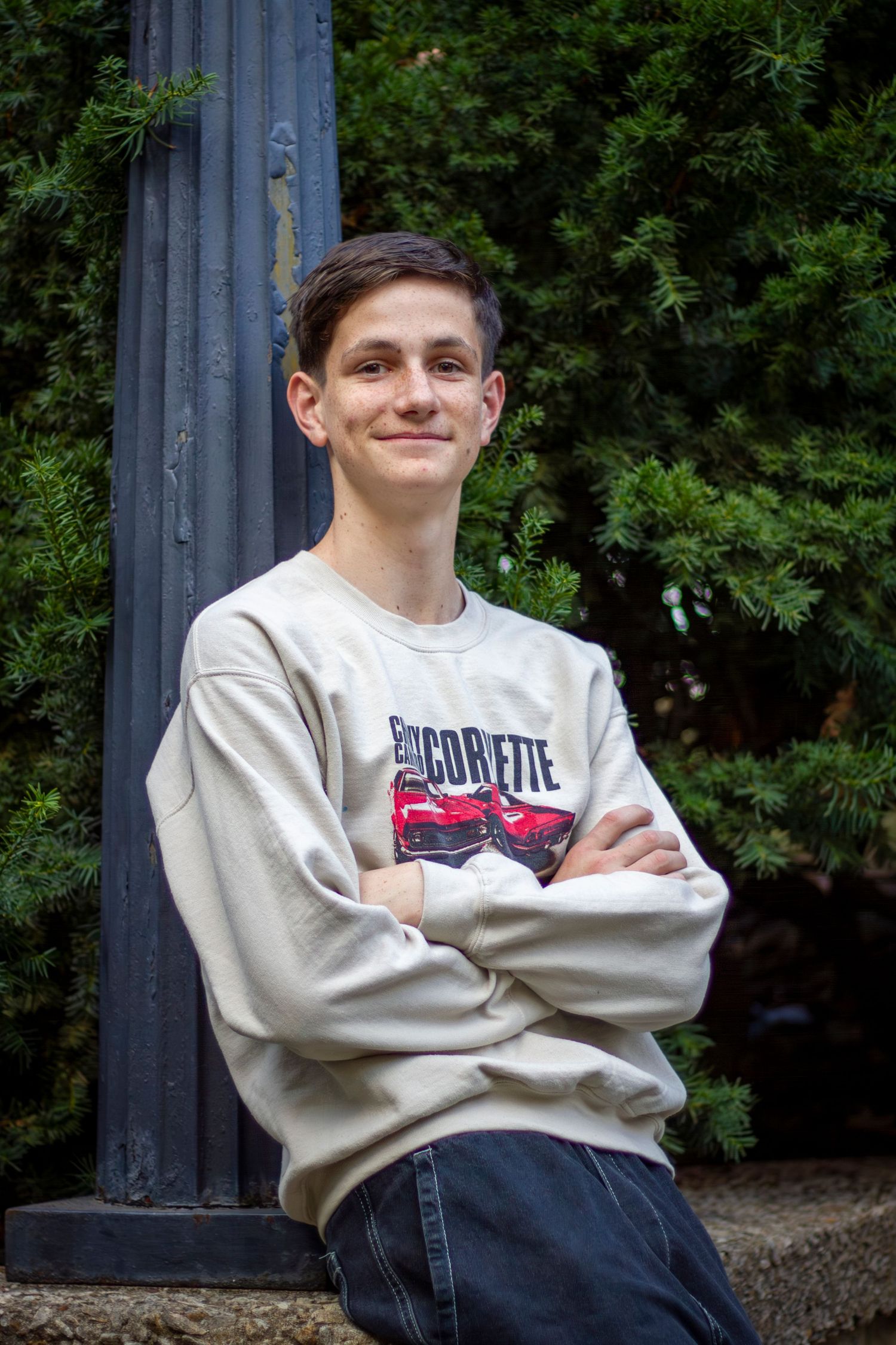 A smiling person in a light-colored sweatshirt sits against a column with trees in the background.