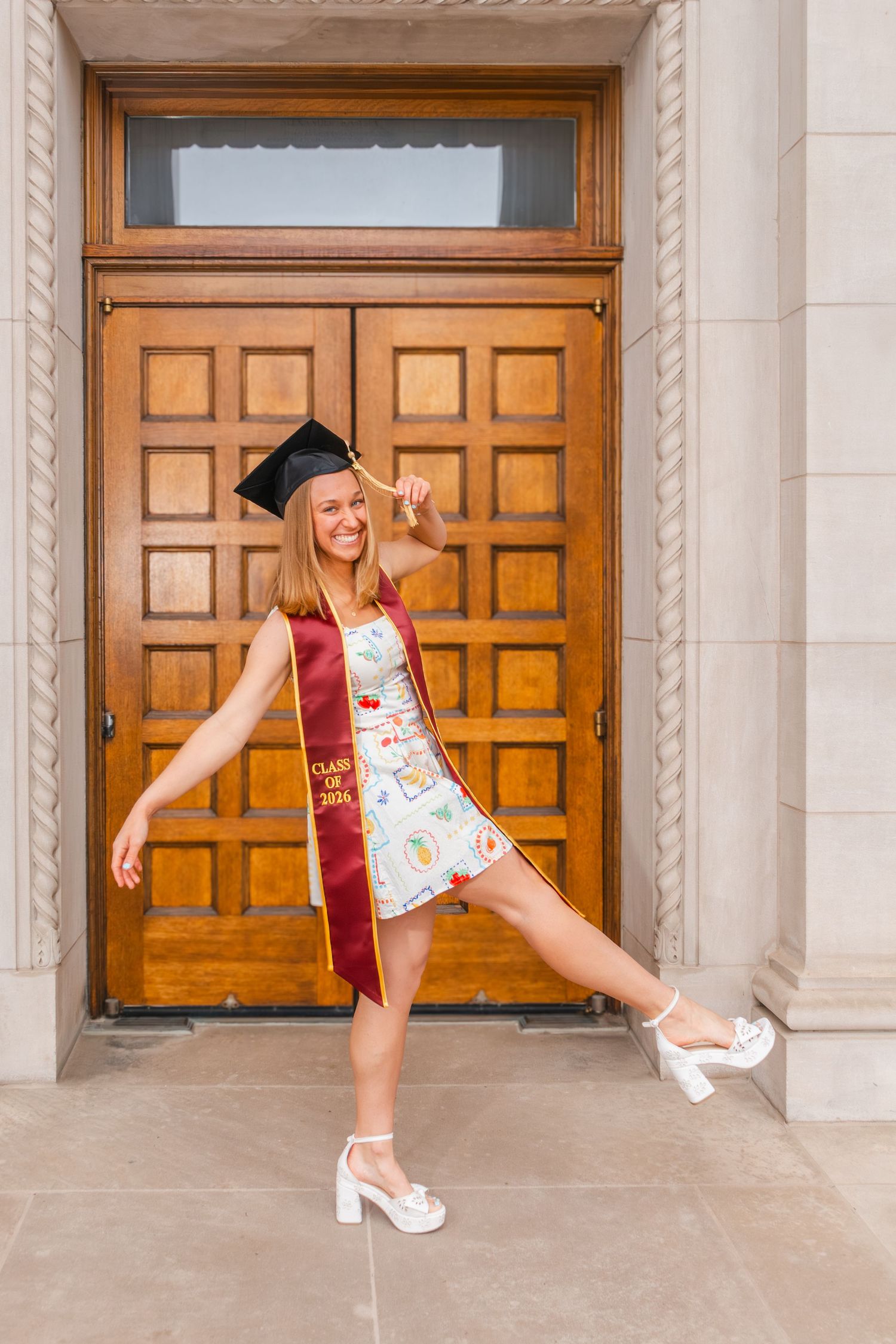 Smiling graduate in cap, sash, and white dress poses playfully in front of wooden doors.