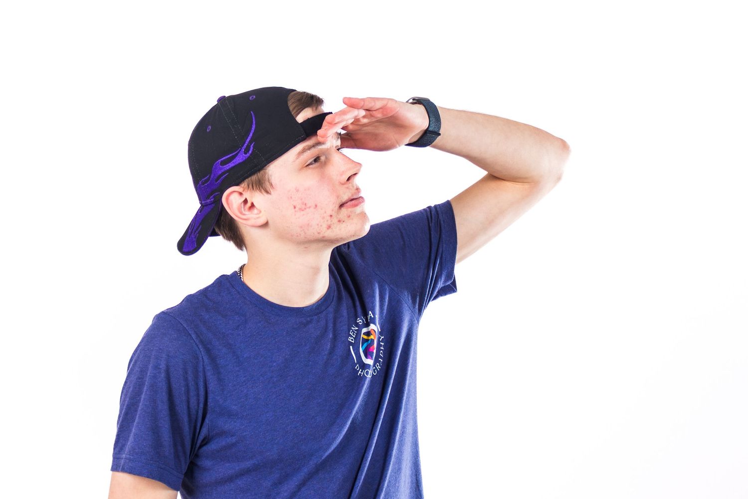Young man in blue t-shirt and black snapback cap shielding eyes with hand against white background.