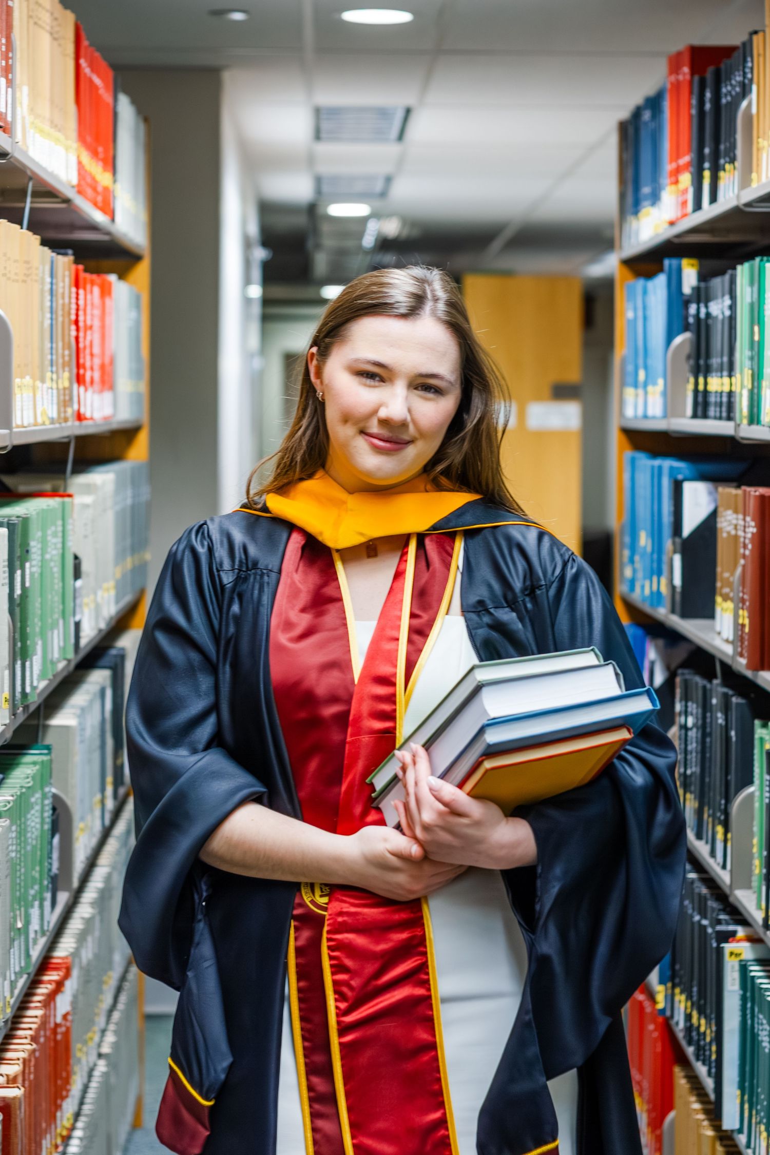 A smiling graduate in cap and gown holds books in a library aisle, wearing gold and red academic regalia.