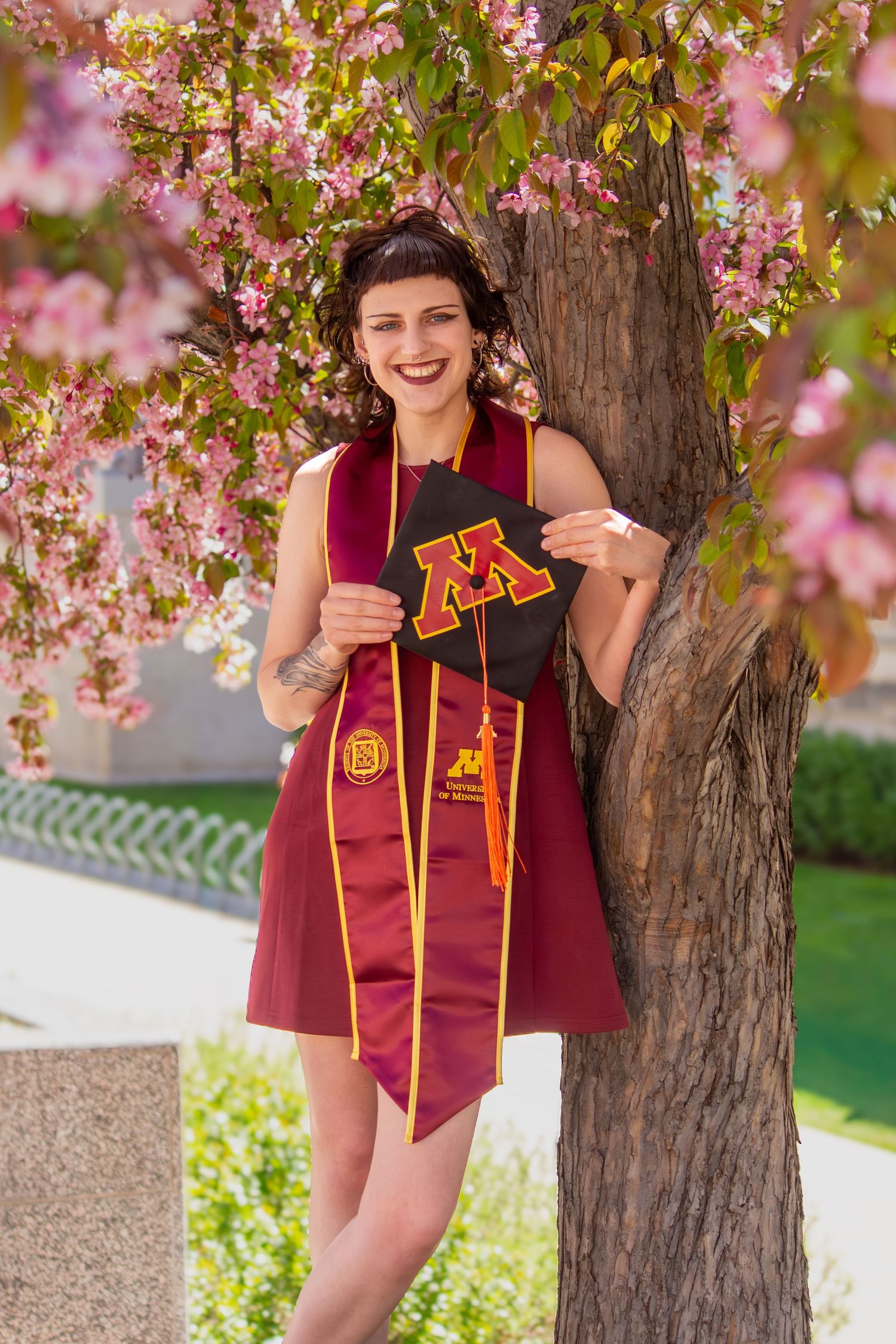 University of Minnesota graduate poses with maroon cap and stole under blooming pink cherry blossom tree in spring.