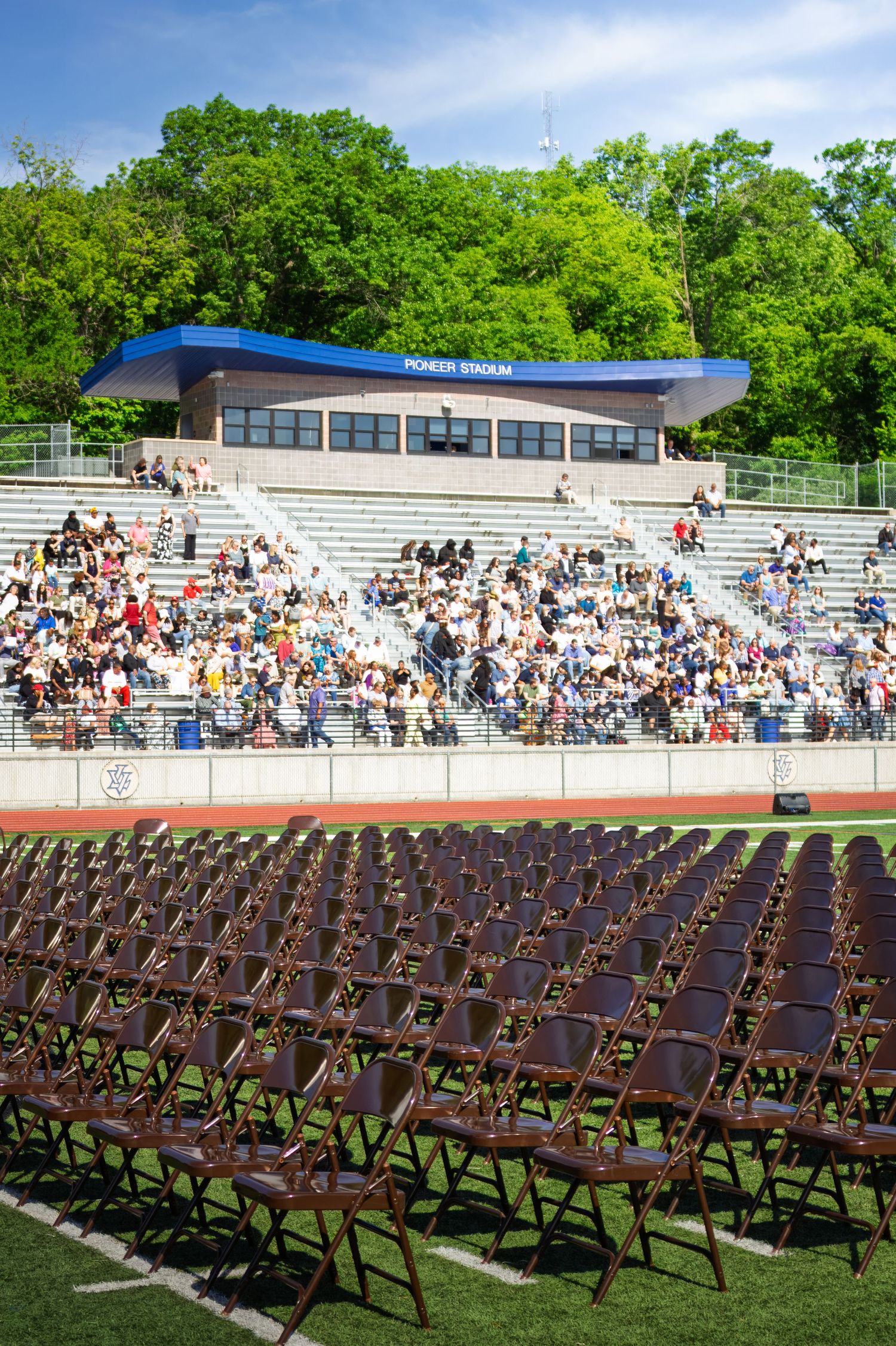 Panoramic view of outdoor stadium bleachers with folding chairs arranged for graduation ceremony beneath trees on sunny day.
