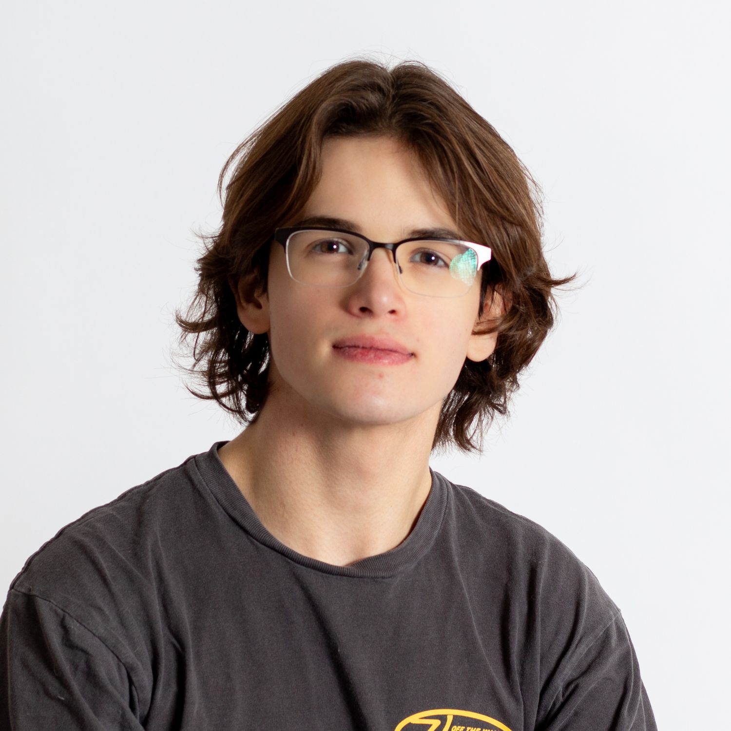 A young person with wavy brown hair wearing glasses and a gray t-shirt poses against a white background.
