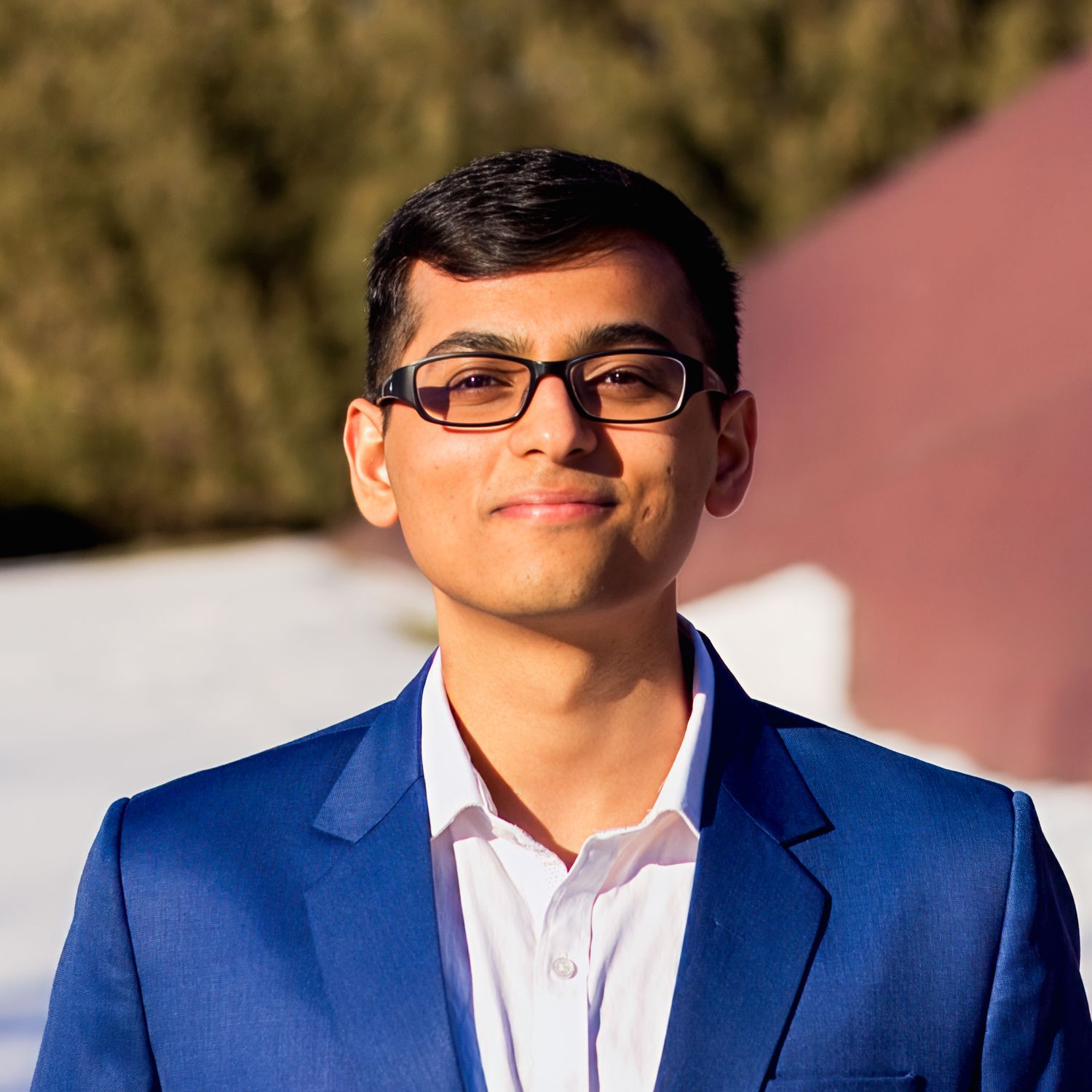 Professional headshot of someone in a navy blazer and white shirt smiling outdoors on a sunny day.