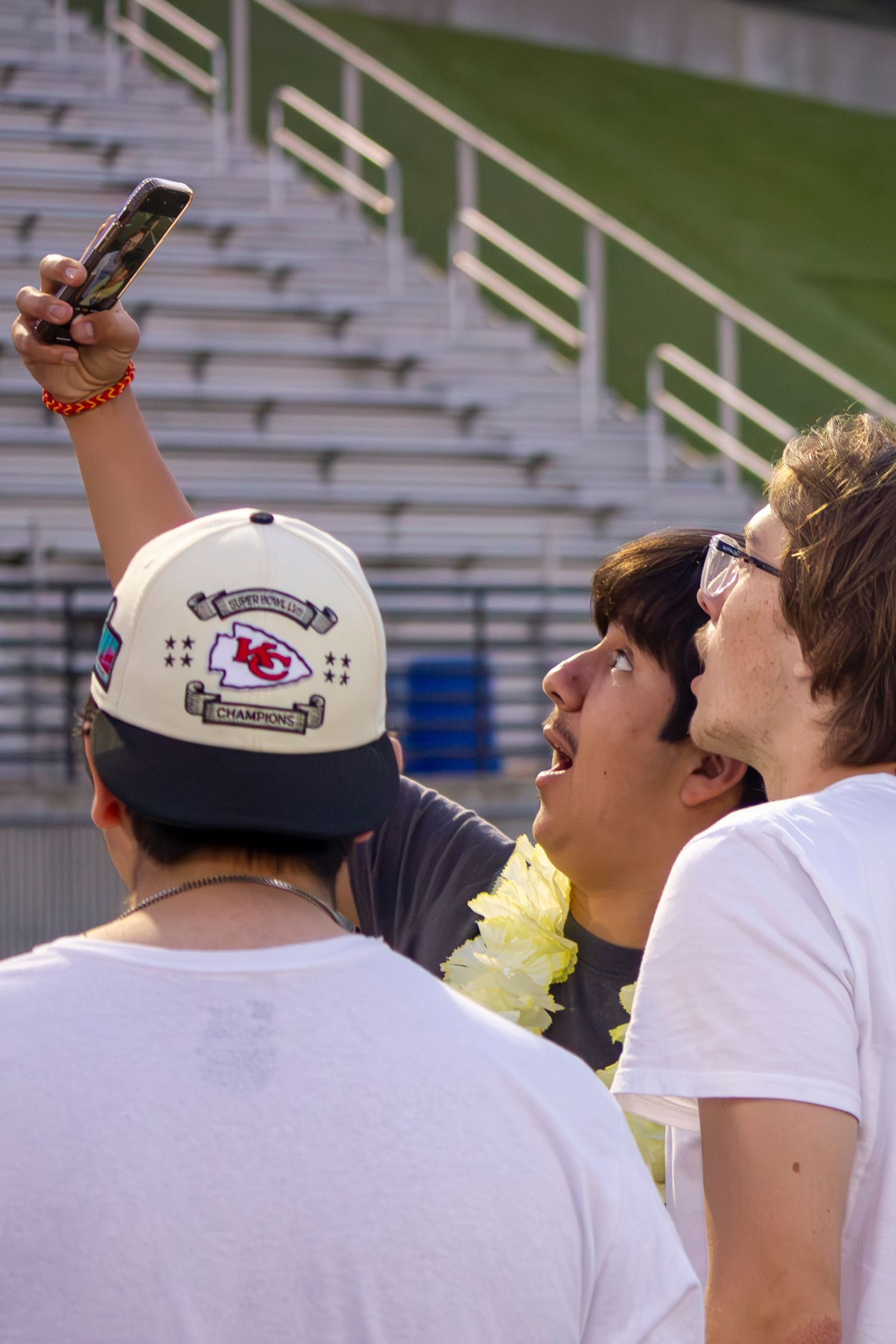 Three friends in casual wear take a selfie in empty stadium seats, one wearing a Kansas City Chiefs baseball cap.