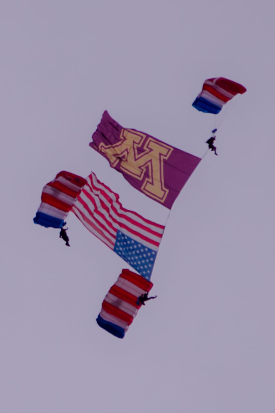 Skydivers with American flags and a University of Minnesota banner performing a formation jump against a purple sky.