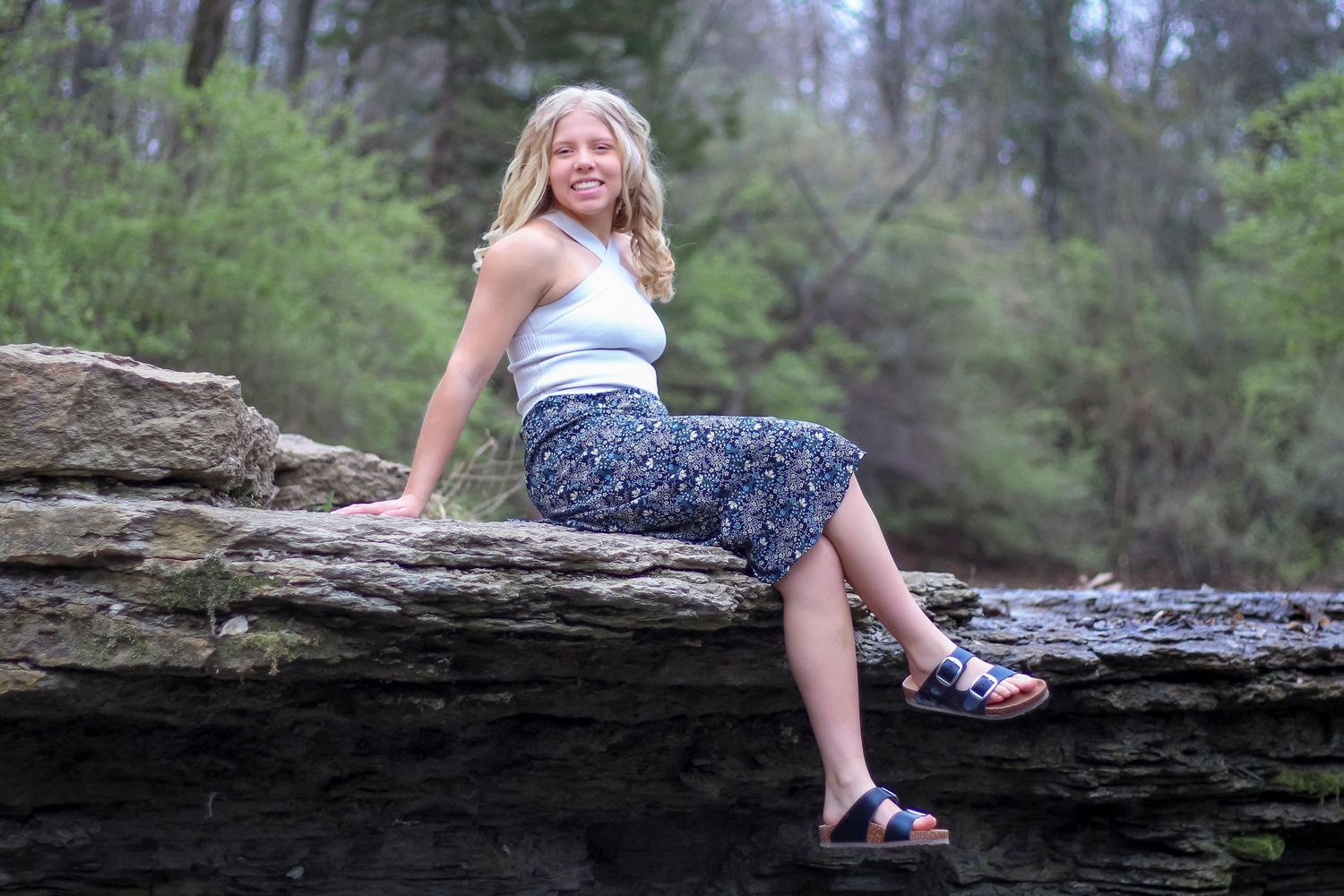 Child in patriotic red white and blue sandals sits on natural stone ledge overlooking wooded landscape.