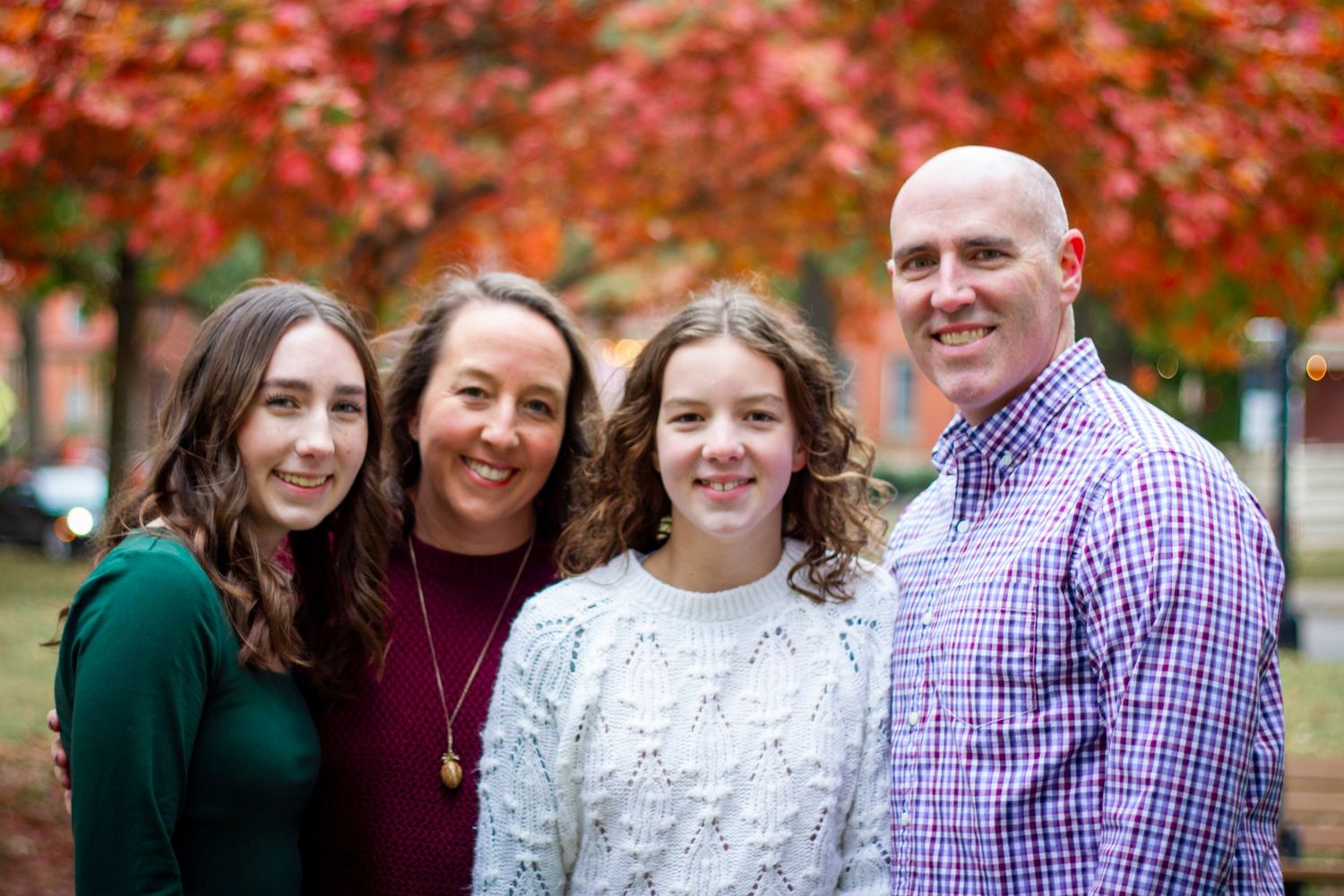 A family of four poses outdoors in autumn with vibrant red maple trees in the background.