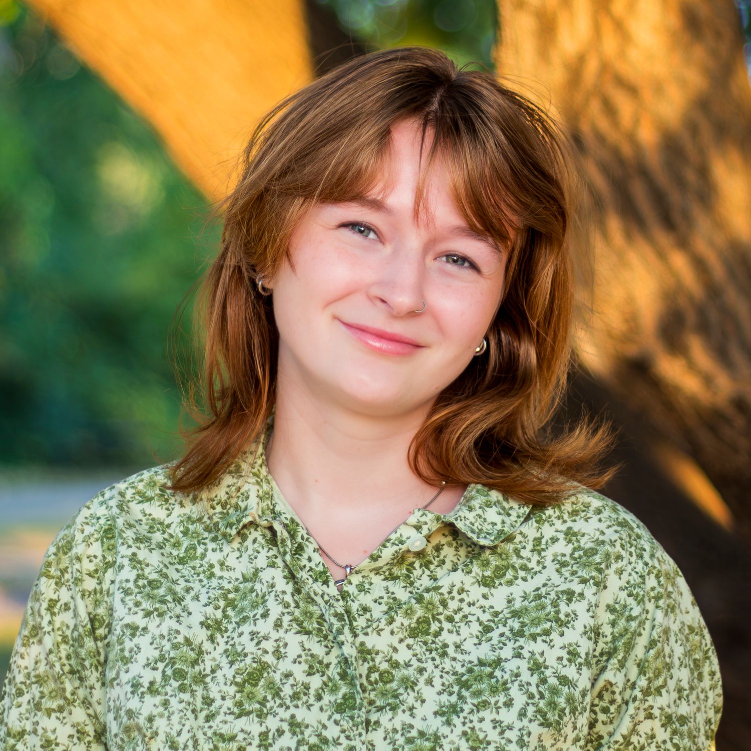 A person in a green floral print top smiles outdoors with warm sunlit trees in the background.