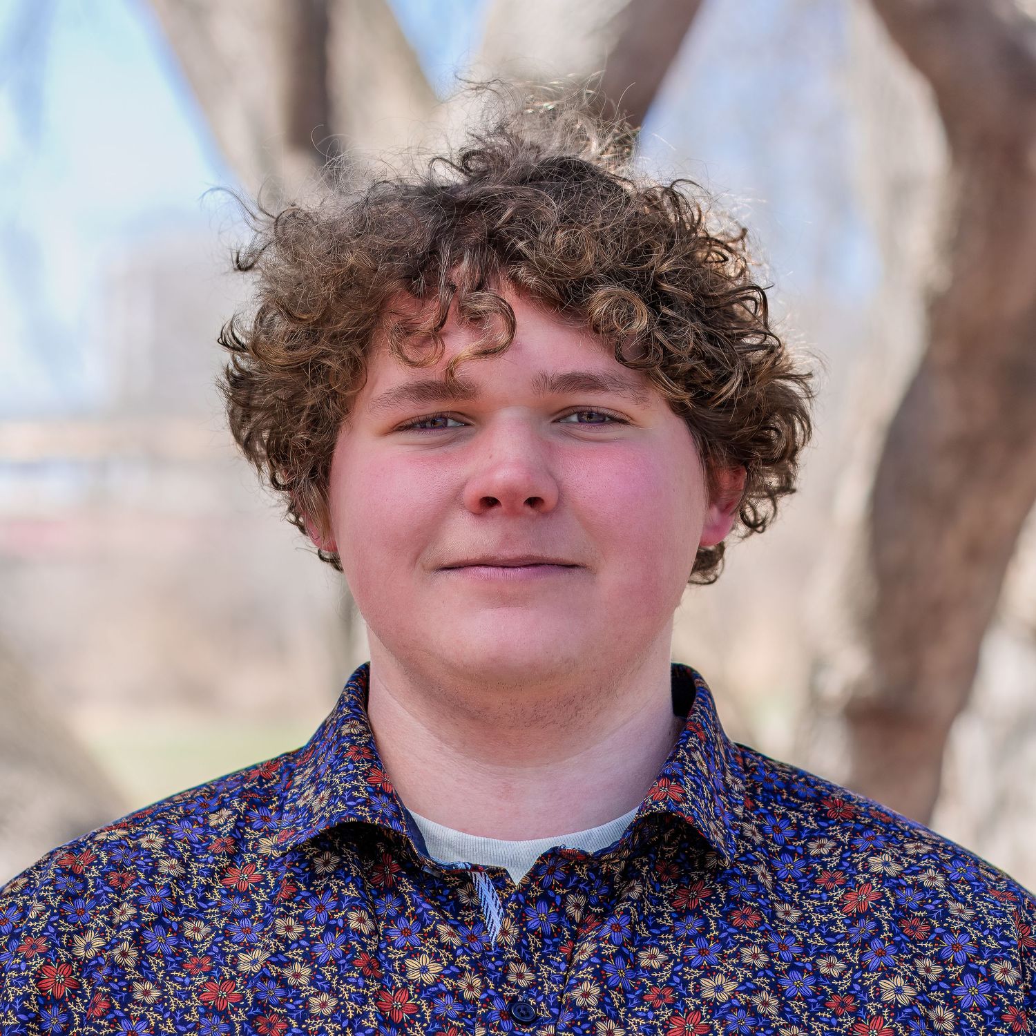 Outdoor portrait of a young person with curly brown hair wearing a blue floral print button-up shirt.