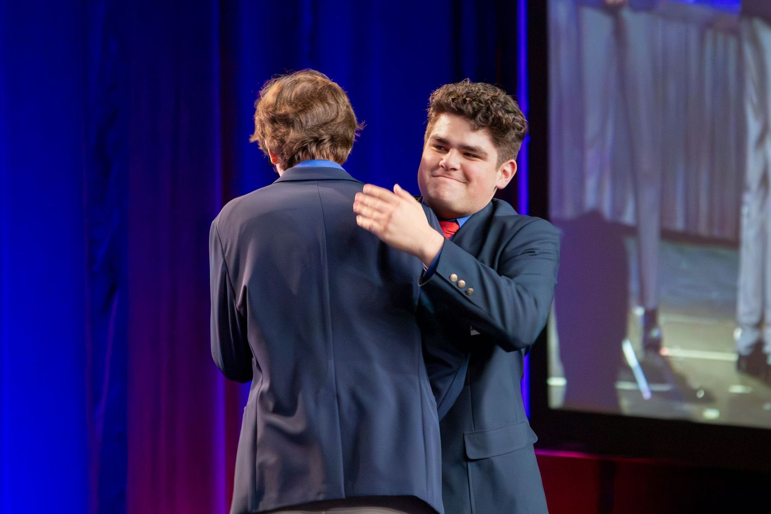 Two people in suits embrace on stage, surrounded by blue lighting.