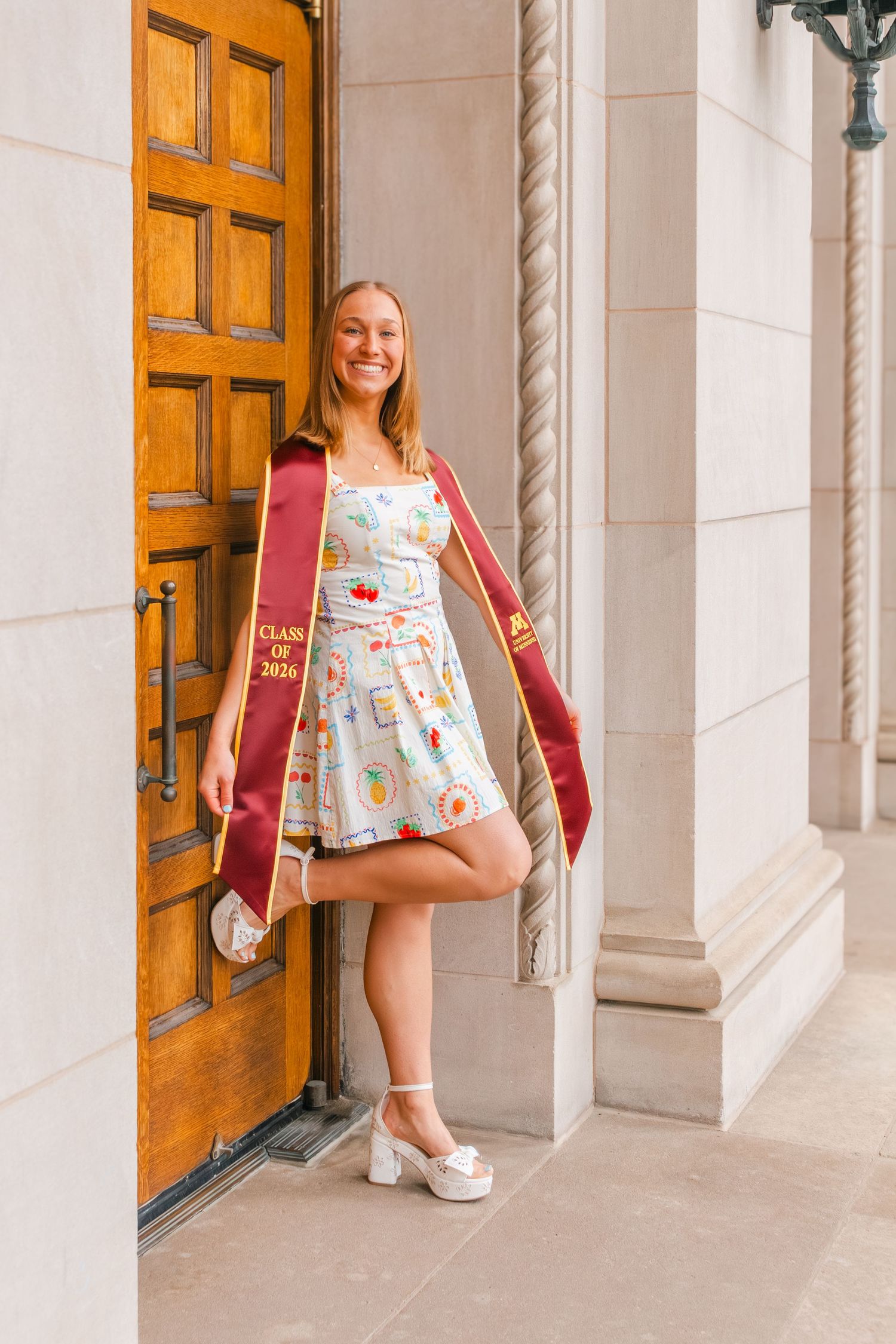 Smiling graduate in floral dress and maroon Class of 2024 sash poses by ornate wooden door between stone columns.