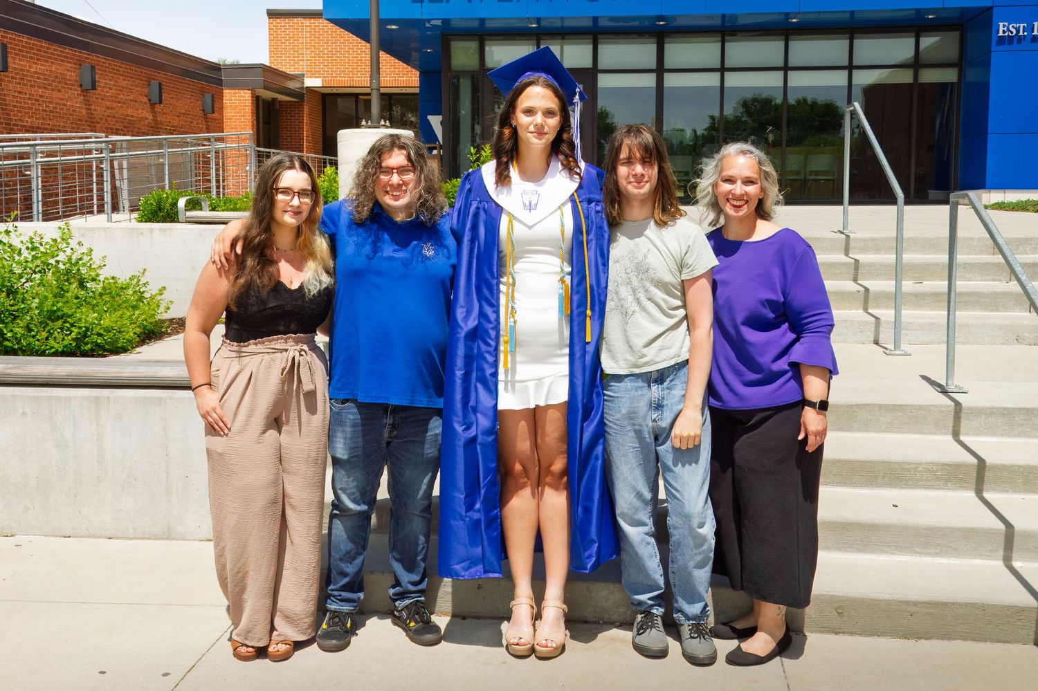 Graduate in blue cap and gown poses on steps outside a building with family members.