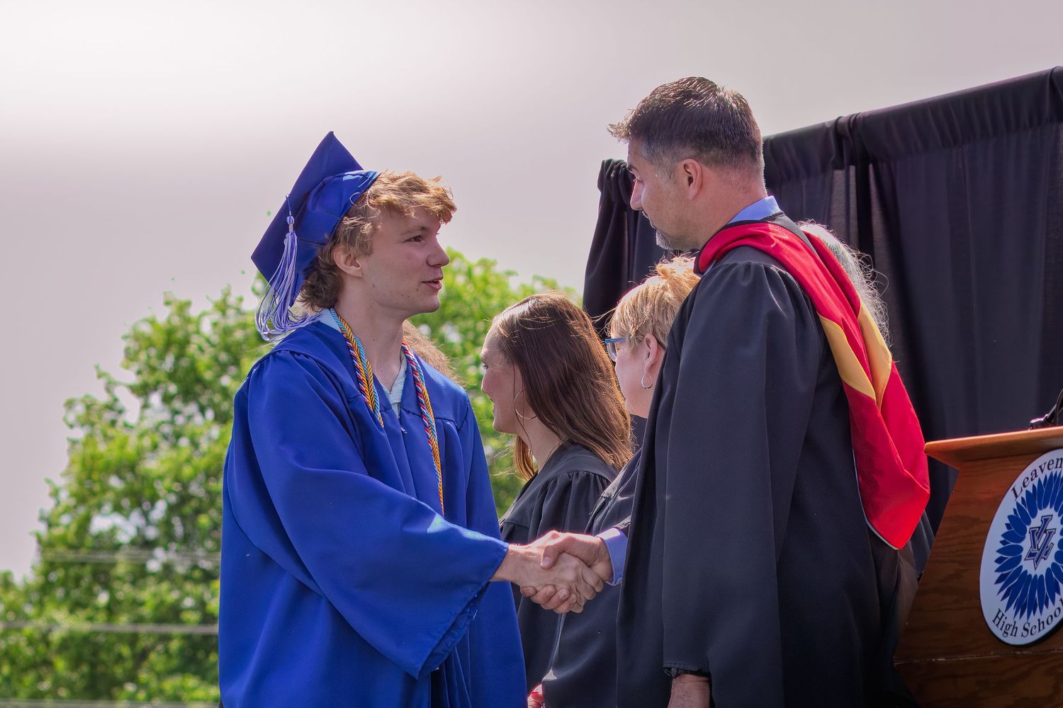 A graduate in bright blue cap and gown shakes hands with faculty member in red and black regalia during commencement ceremony.