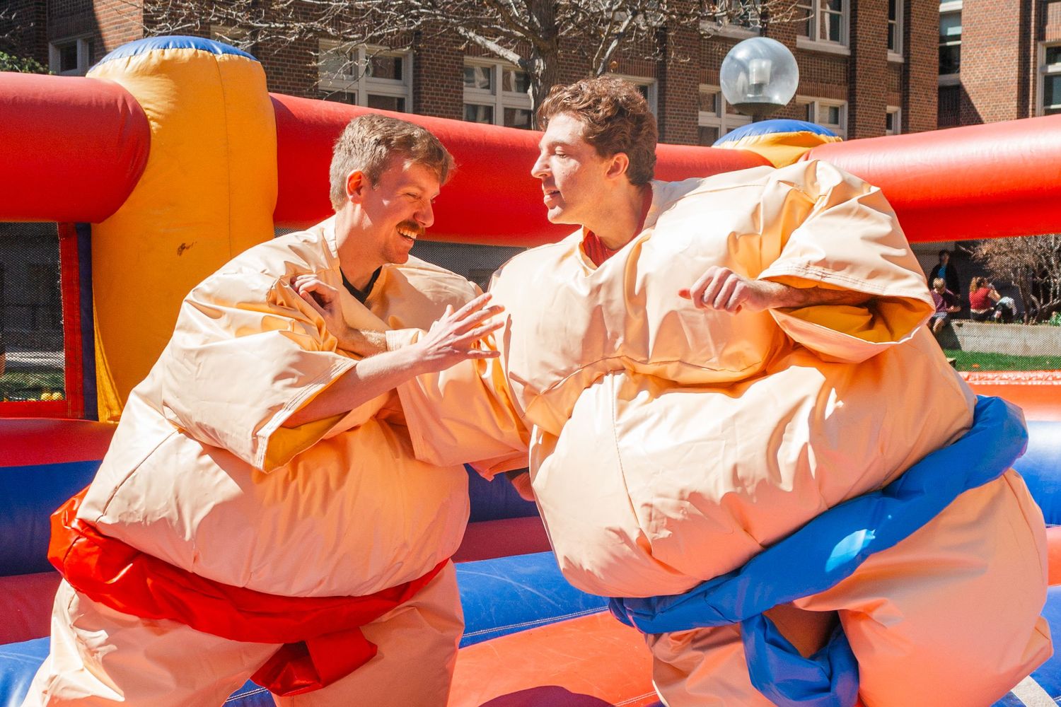 Two young men wearing inflatable sumo wrestler suits compete in a colorful bouncy arena outdoors.
