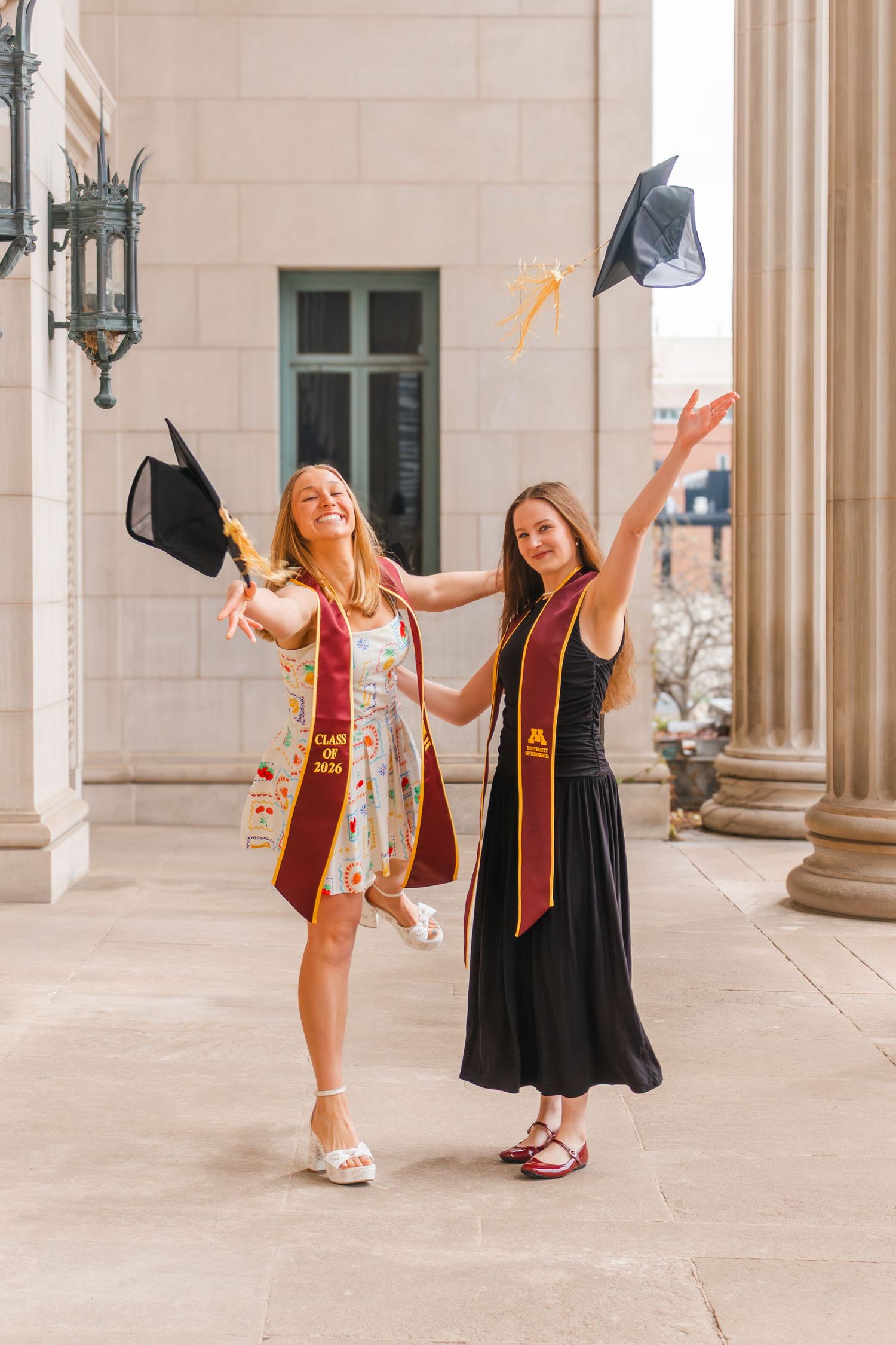 Two smiling graduates in maroon stoles joyfully toss their caps in the air outside a columned building.