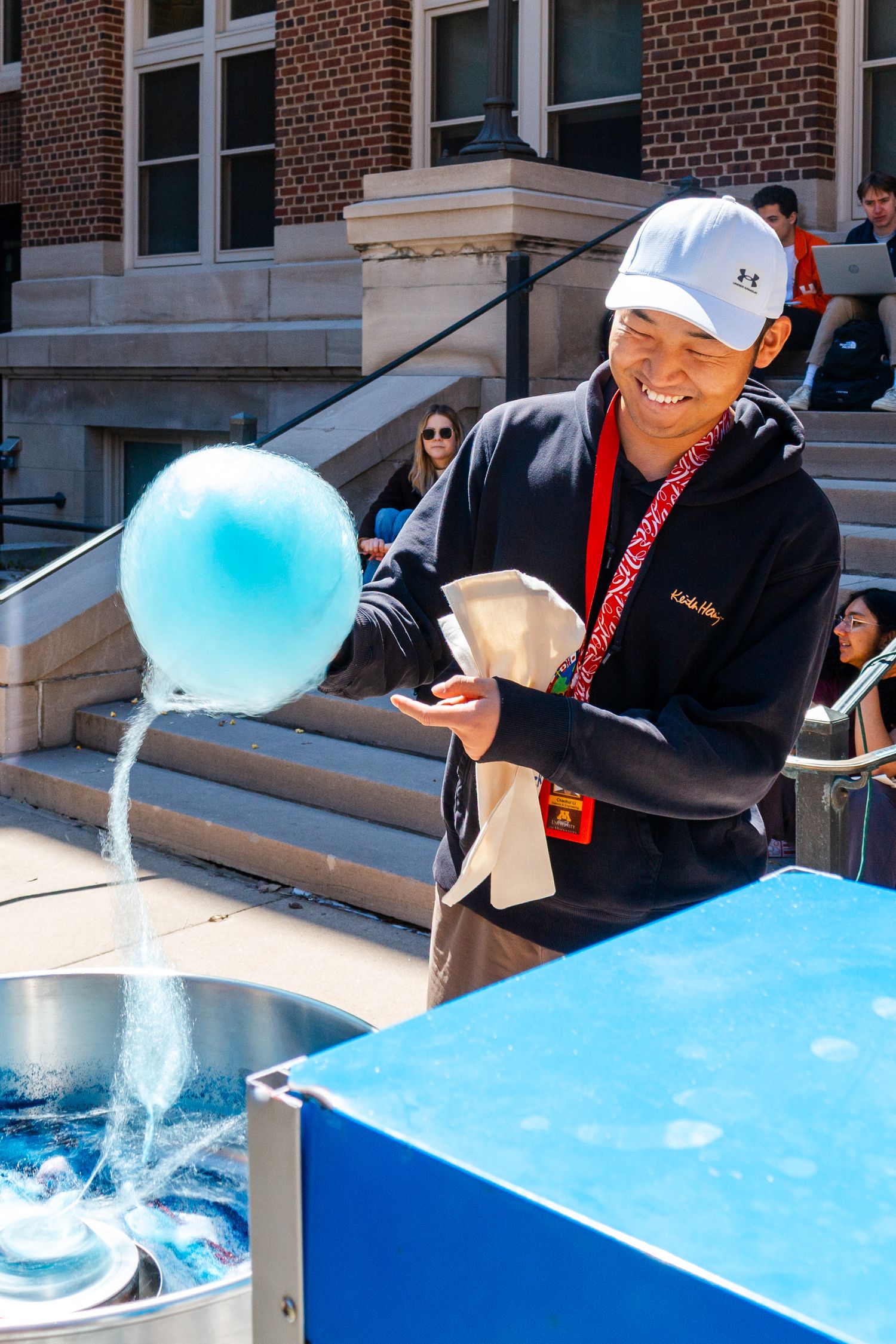 Man in white cap smiling while making blue cotton candy at an outdoor event on a sunny day.