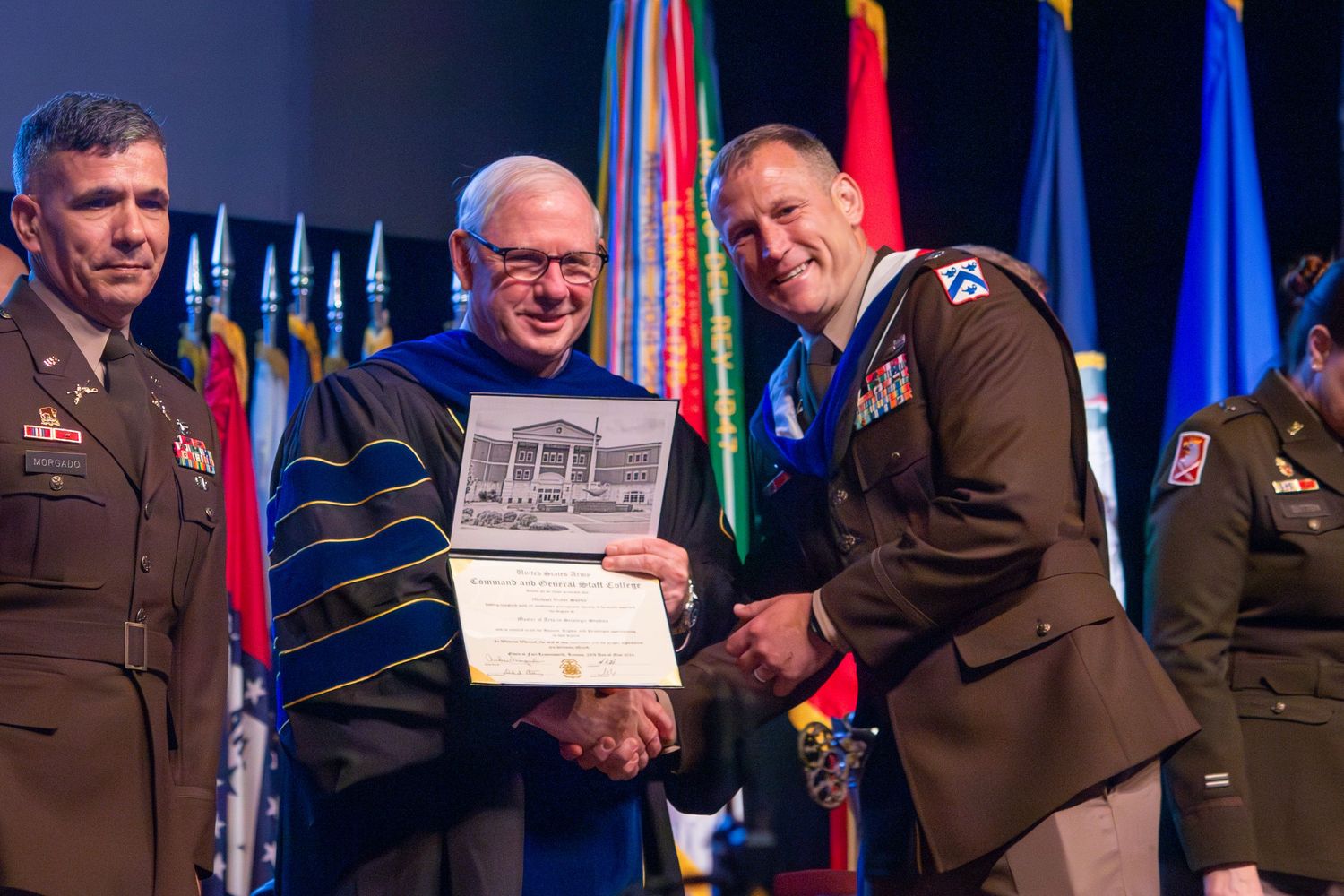 Military officers and academic official in ceremonial dress present and receive awards on stage with flags in background.