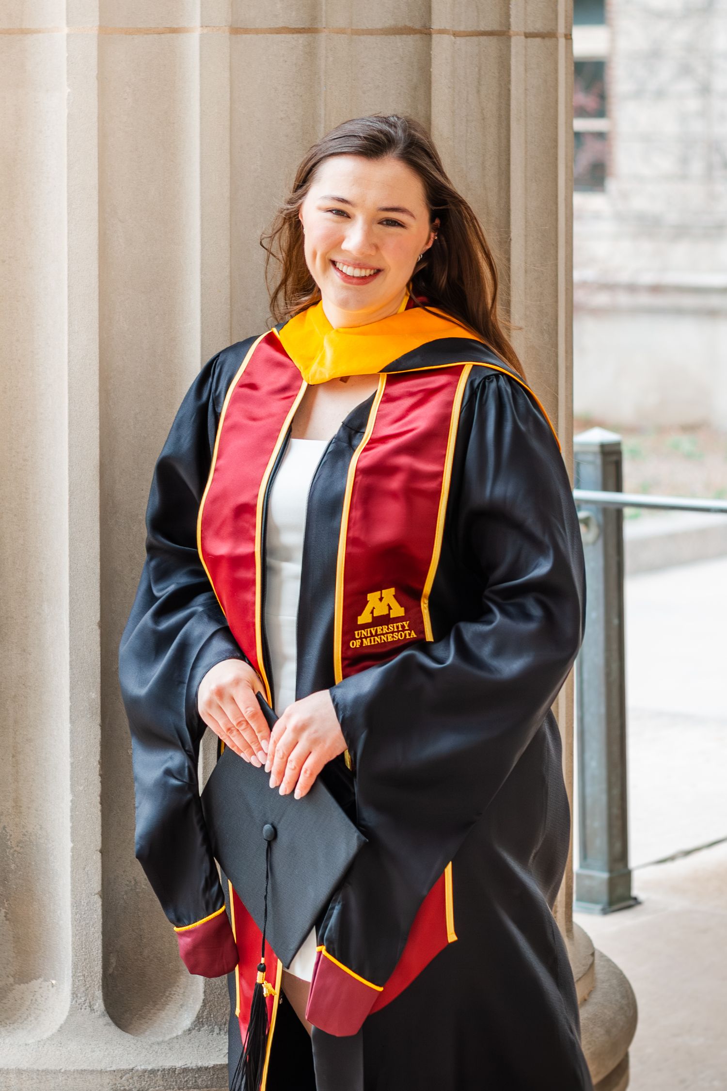 Smiling young woman in University of Minnesota graduation gown with maroon and gold regalia posing by stone columns.