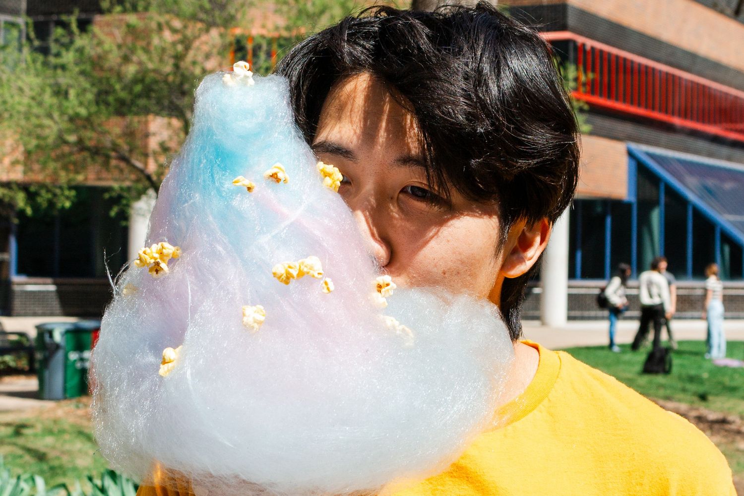 Young man in yellow shirt eating large blue cotton candy with popcorn toppings outdoors on sunny day.