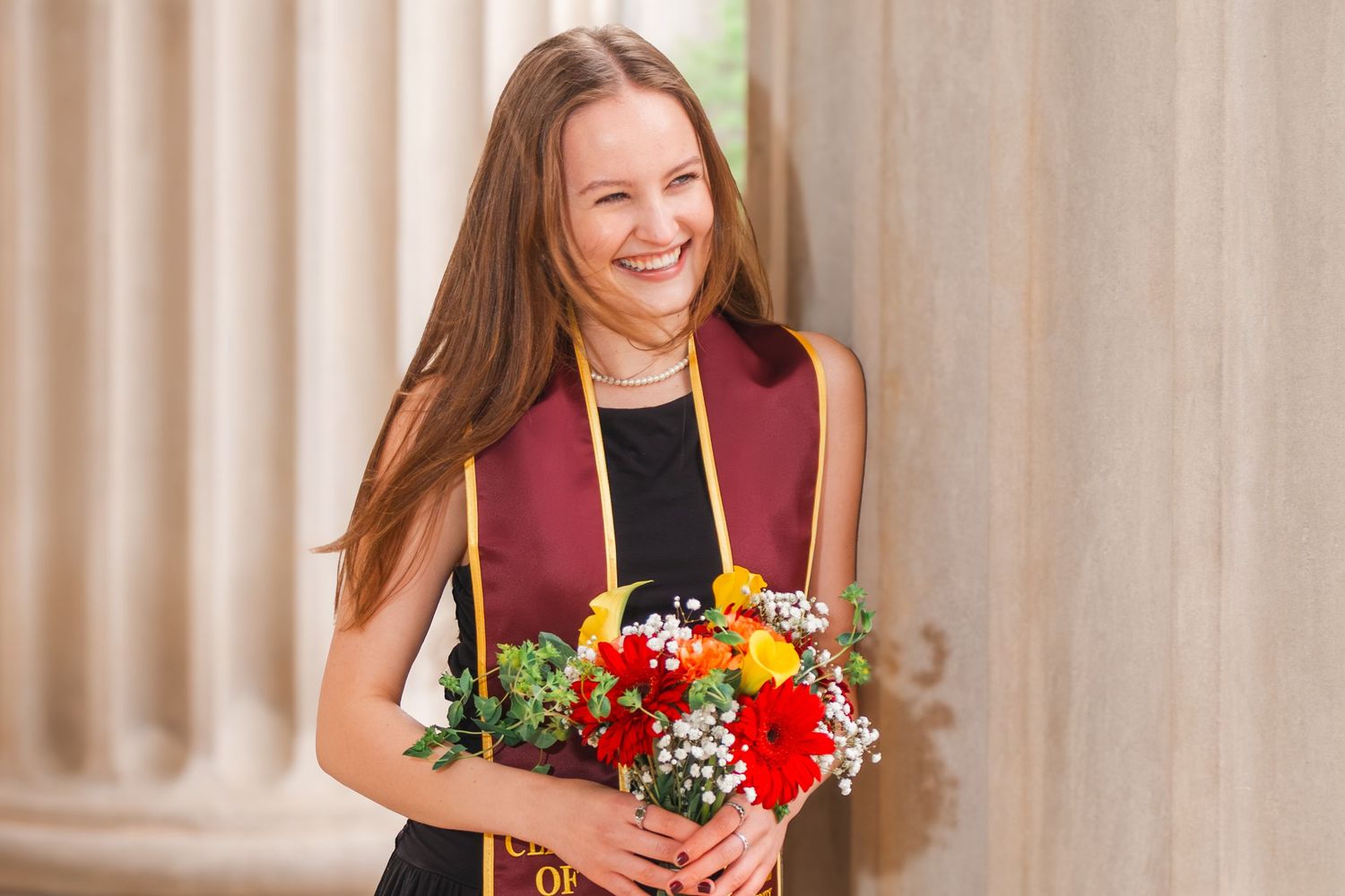Smiling young woman in graduation regalia holding colorful bouquet of flowers near stone columns.