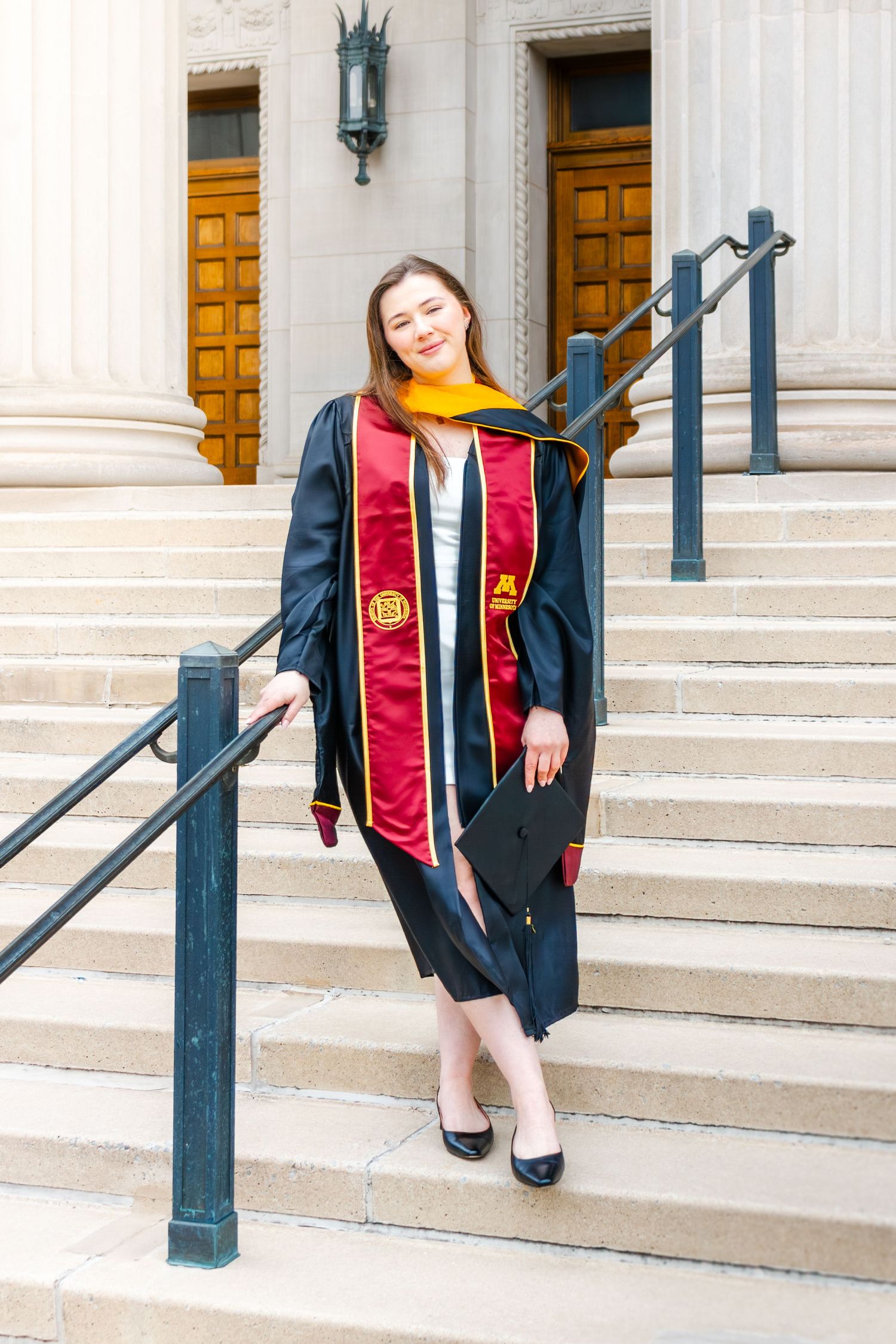 Young woman in black graduation gown with red and gold stole poses on university steps.