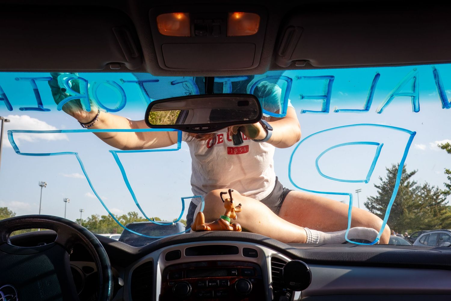 A graduate paints her name on a car windshield, shown from inside the car.