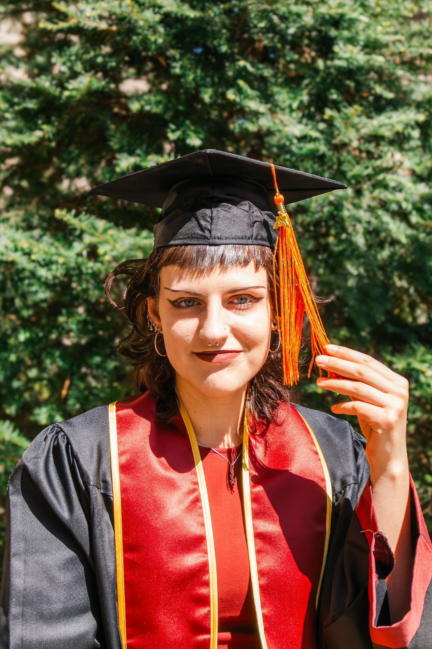 Young woman in black and red graduation gown and cap holding orange tassel, smiling outdoors.